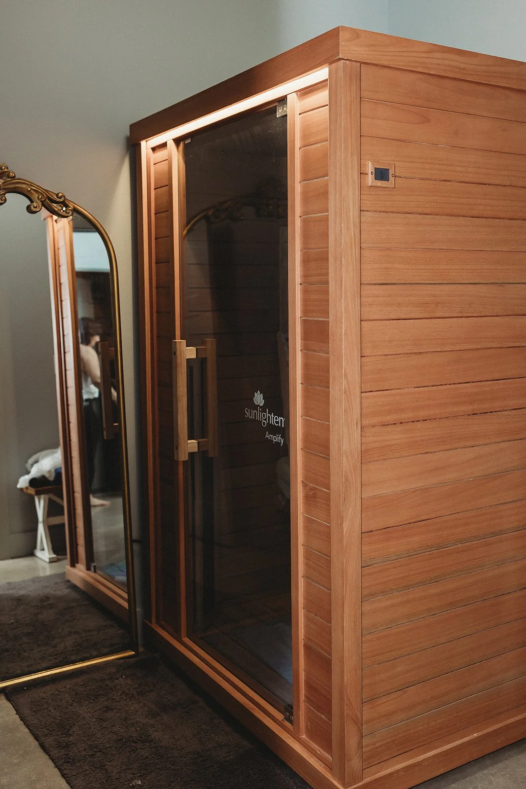 A wooden infrared sauna with a glass door, situated next to a mirror in a room with a brown rug and a person visible in the mirror's reflection.