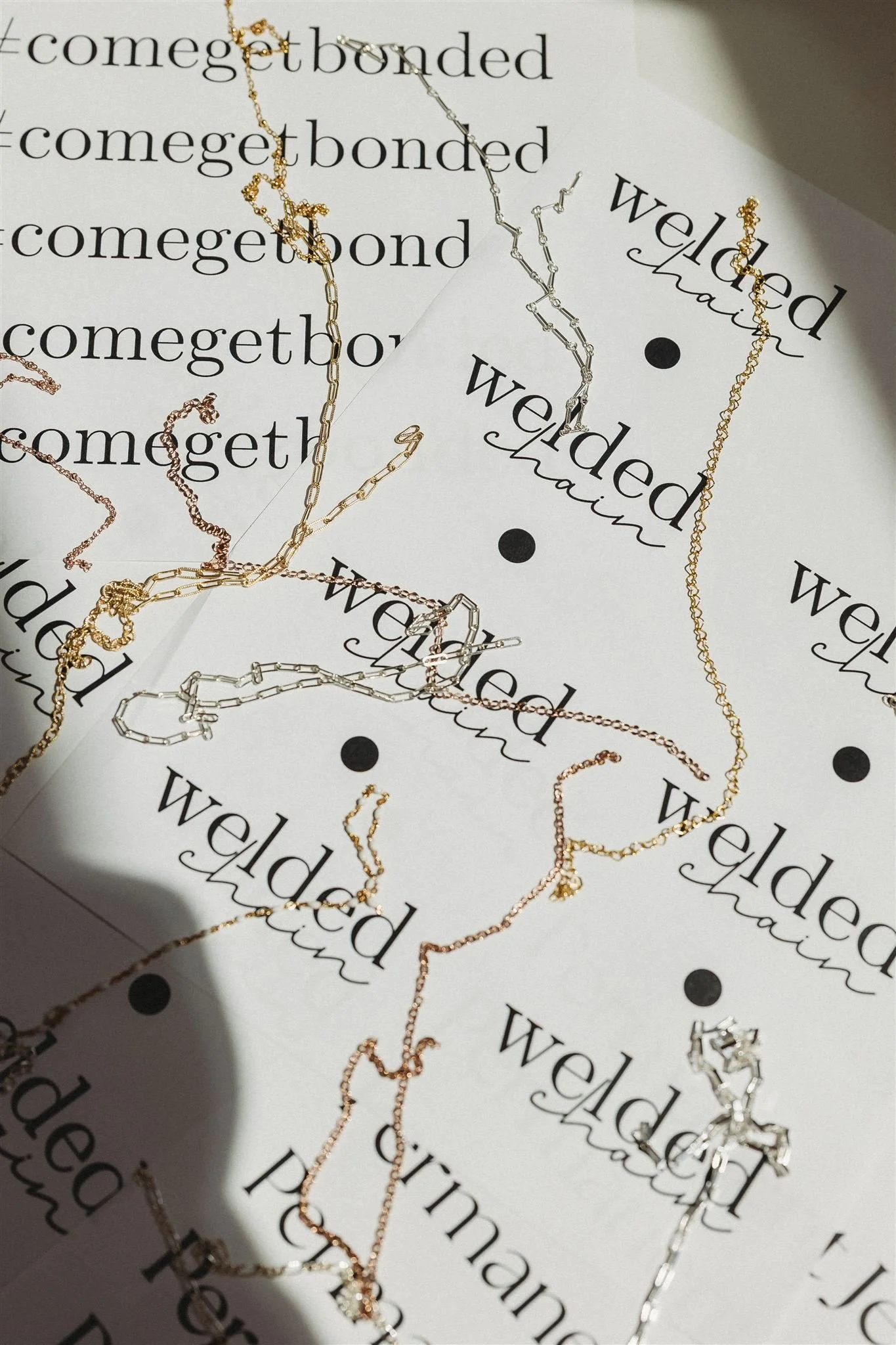 Multiple delicate gold and silver necklaces are arranged on a paper background that has the repeated phrase 'welded chain' printed on it.