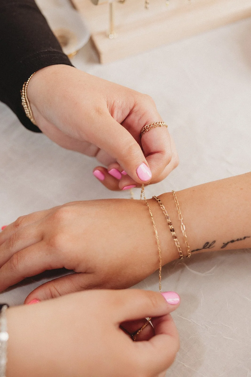 Close-up of a person's wrist with jewelry, as another person adds a gold bracelet. The person has pink nail polish and a tattoo on the inner wrist, with a jewelry display in the background.