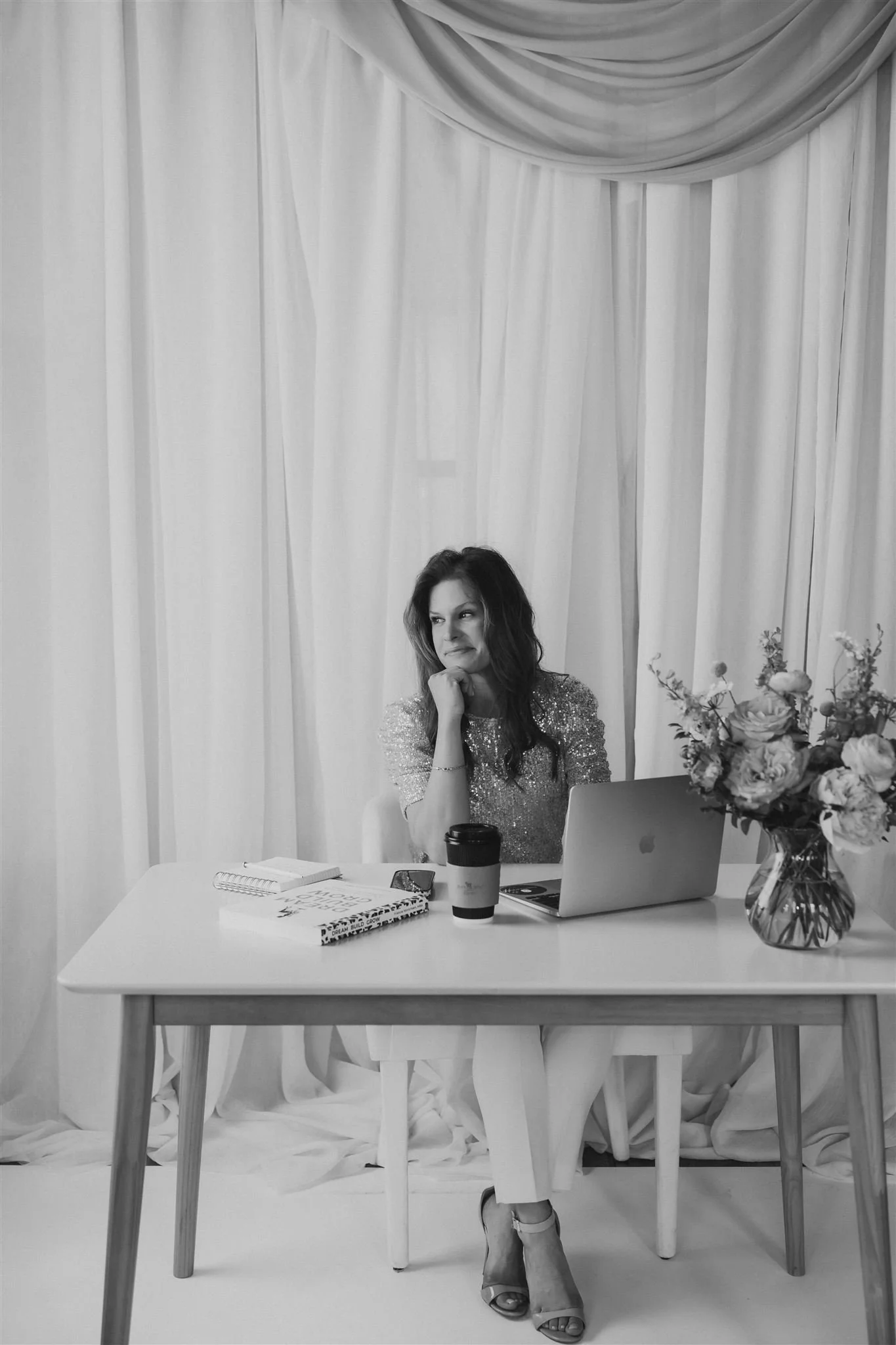 A woman sitting at a desk with a laptop, notebooks, a coffee cup, and a vase of flowers, in front of white curtains.