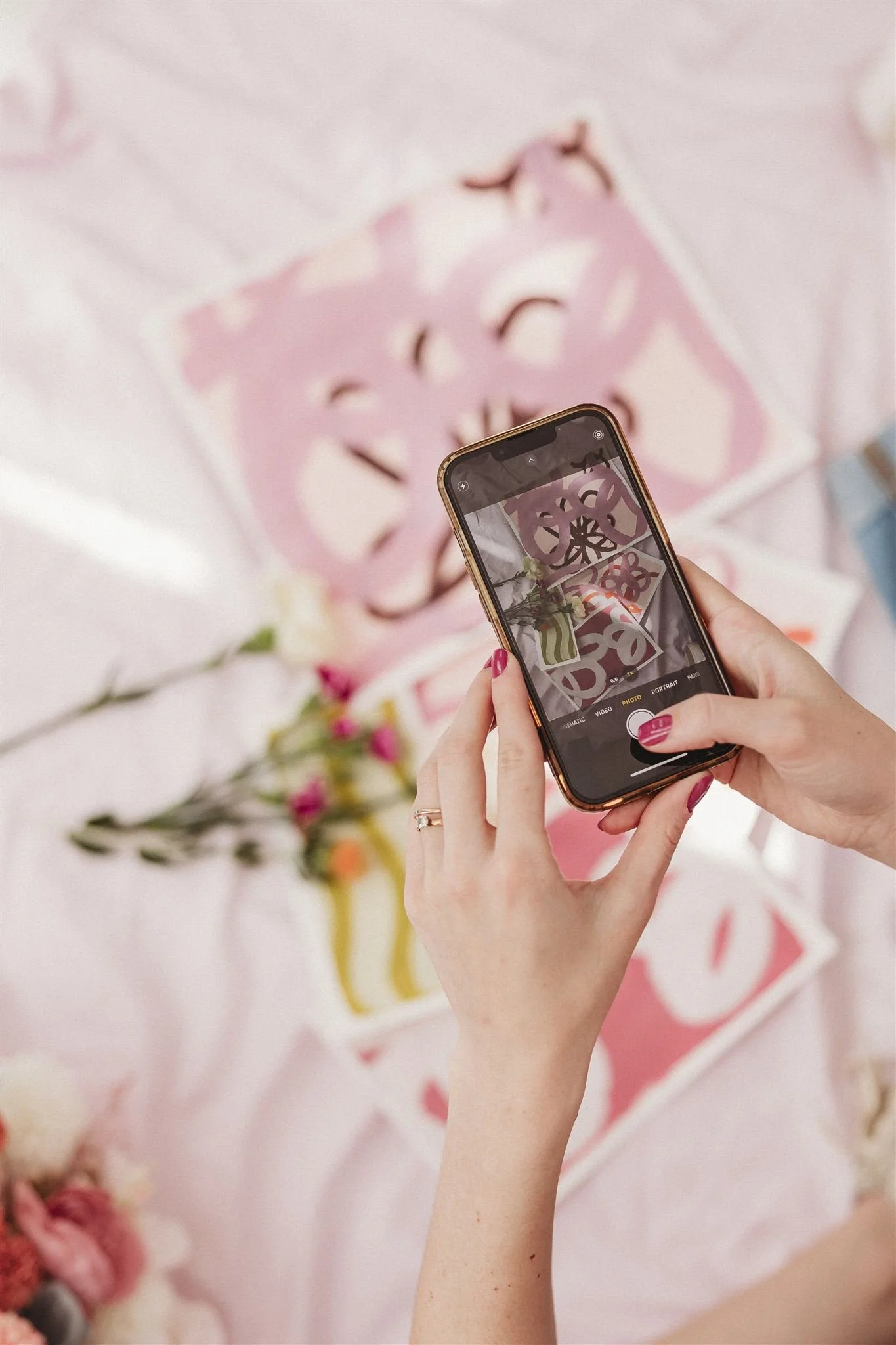 Person taking a photo of a pink decorative item with a smartphone, with flowers and a pink cloth background.