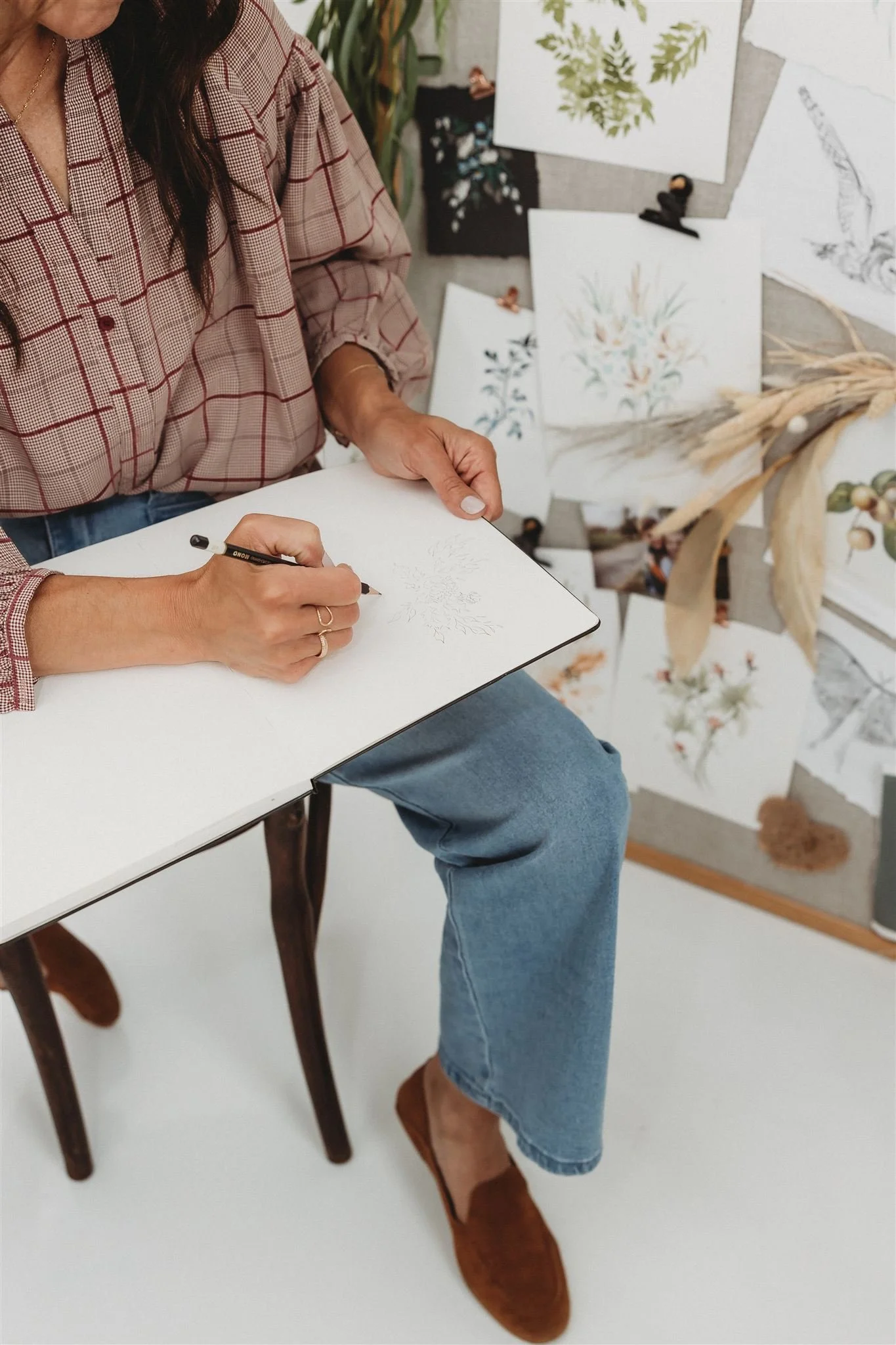 Person drawing on a sketchpad with botanical illustrations on a wall in the background.