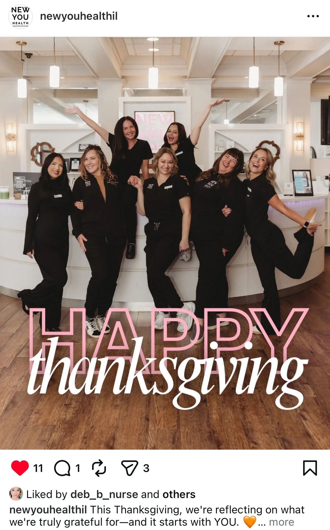 A group of seven women wearing black scrubs, standing behind a curved reception desk, posing happily with arms raised and around each other in a professional setting. The background shows a well-lit, modern interior with hanging lights and framed dec
