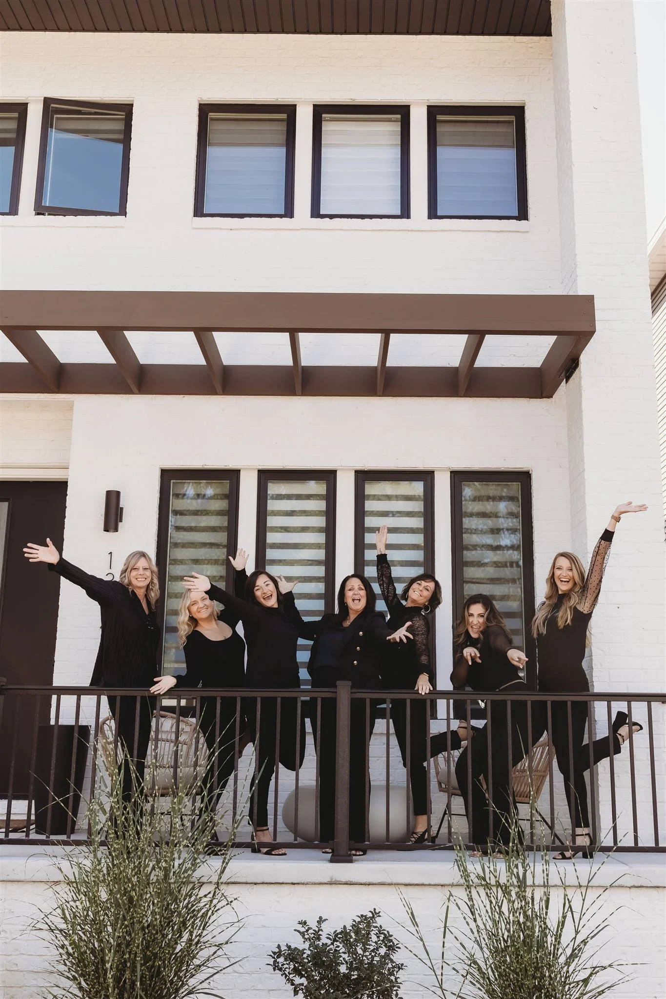 Group of seven women in black outfits on a balcony, smiling, and posing cheerfully in front of a white building with large windows.