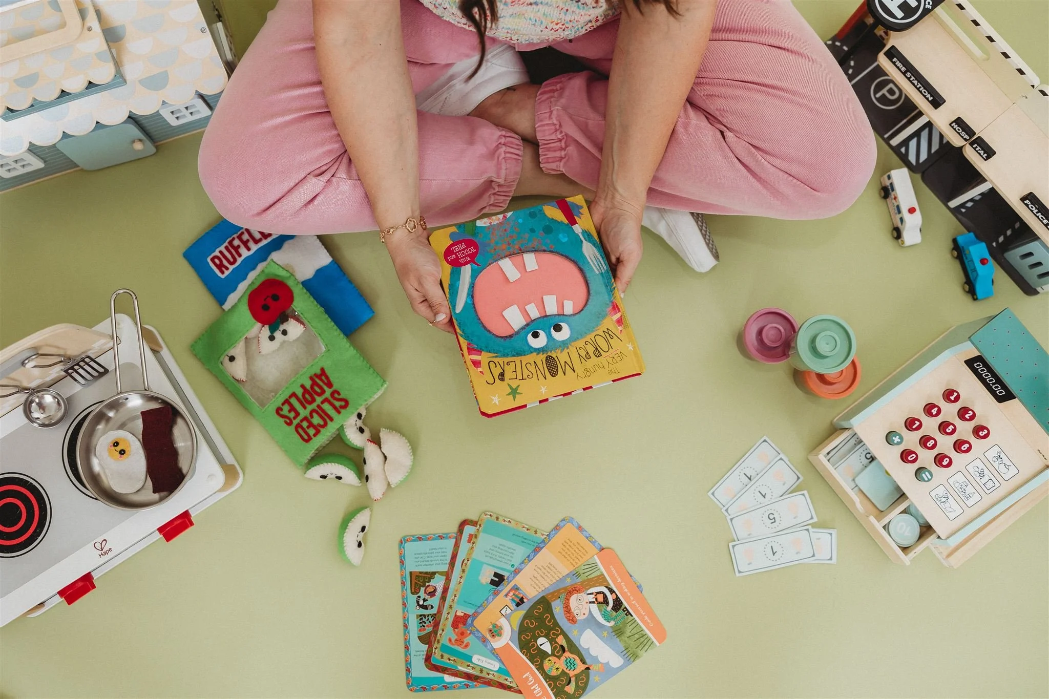 Overhead view of a woman in pink pants sitting cross-legged on a sage green floor, holding a children's book called "Worry Monsters," surrounded by wooden play kitchen toys, felt food, a toy cash register, and illustrated activity cards.