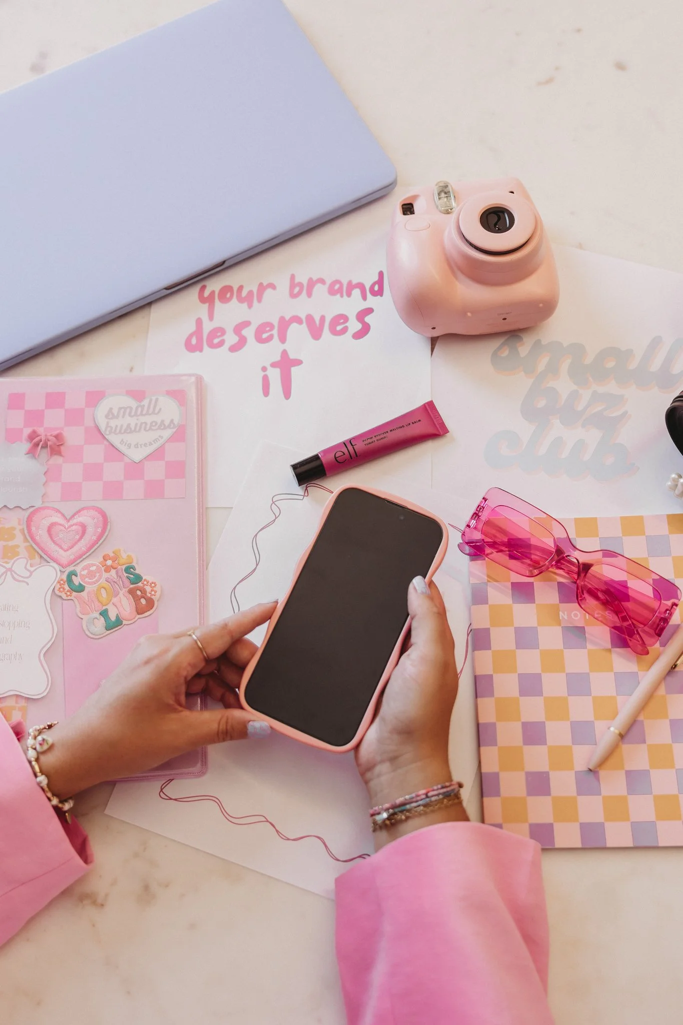 A person in a pink long-sleeve shirt holding a smartphone with a pink case on a white table with various pink-themed items, including a pink camera, pink sunglasses, a pink pen, notebooks, and stickers.