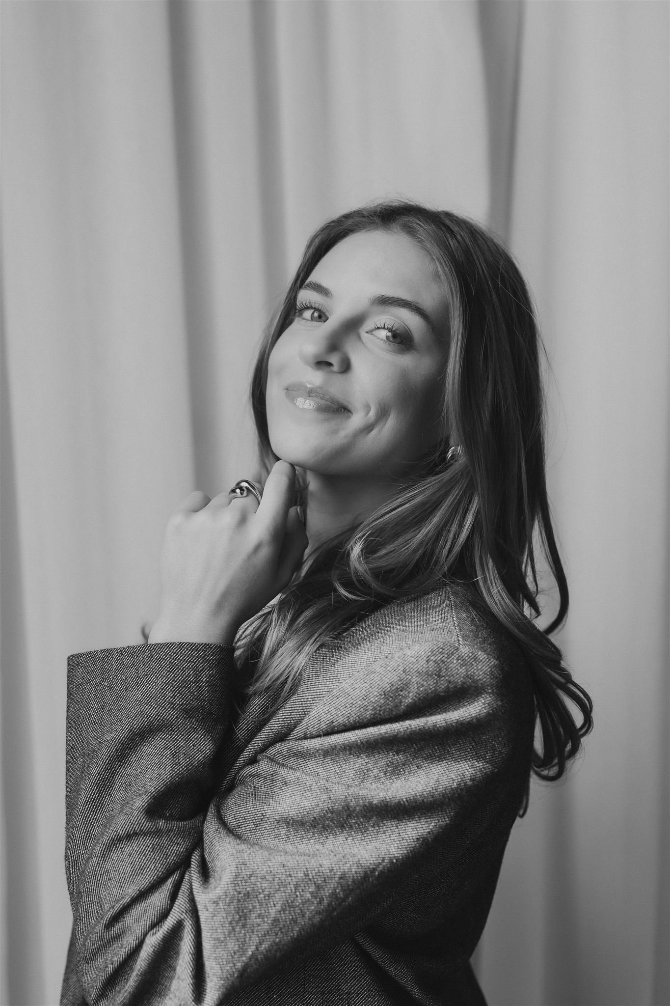 Black and white portrait of a woman with long wavy hair, smiling softly, dressed in a blazer, standing in front of curtains.