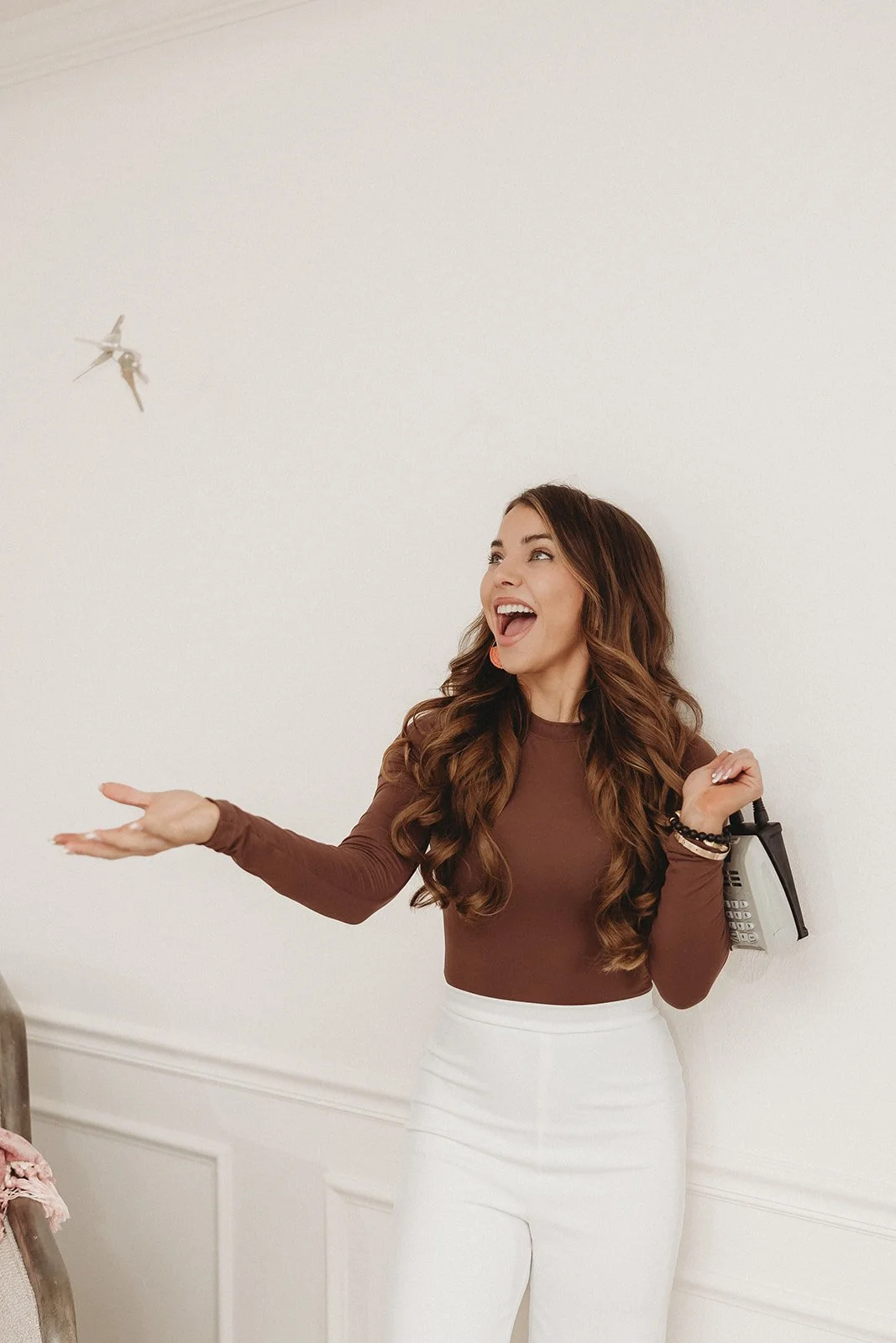 A woman with long wavy brown hair wearing a brown long-sleeve top and white pants, smiling and throwing a paper airplane in a white room.