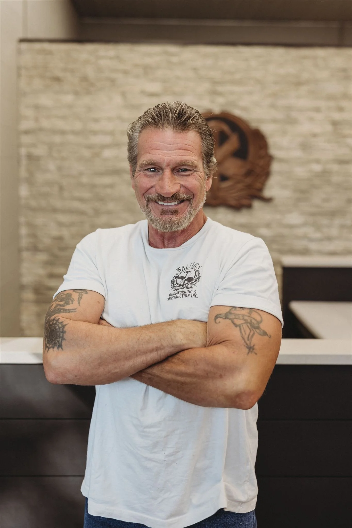 A smiling middle-aged man with Salt and Pepper hair and beard, arms crossed, standing indoors against a beige stone wall with a wooden emblem behind him, wearing a white t-shirt with a logo.