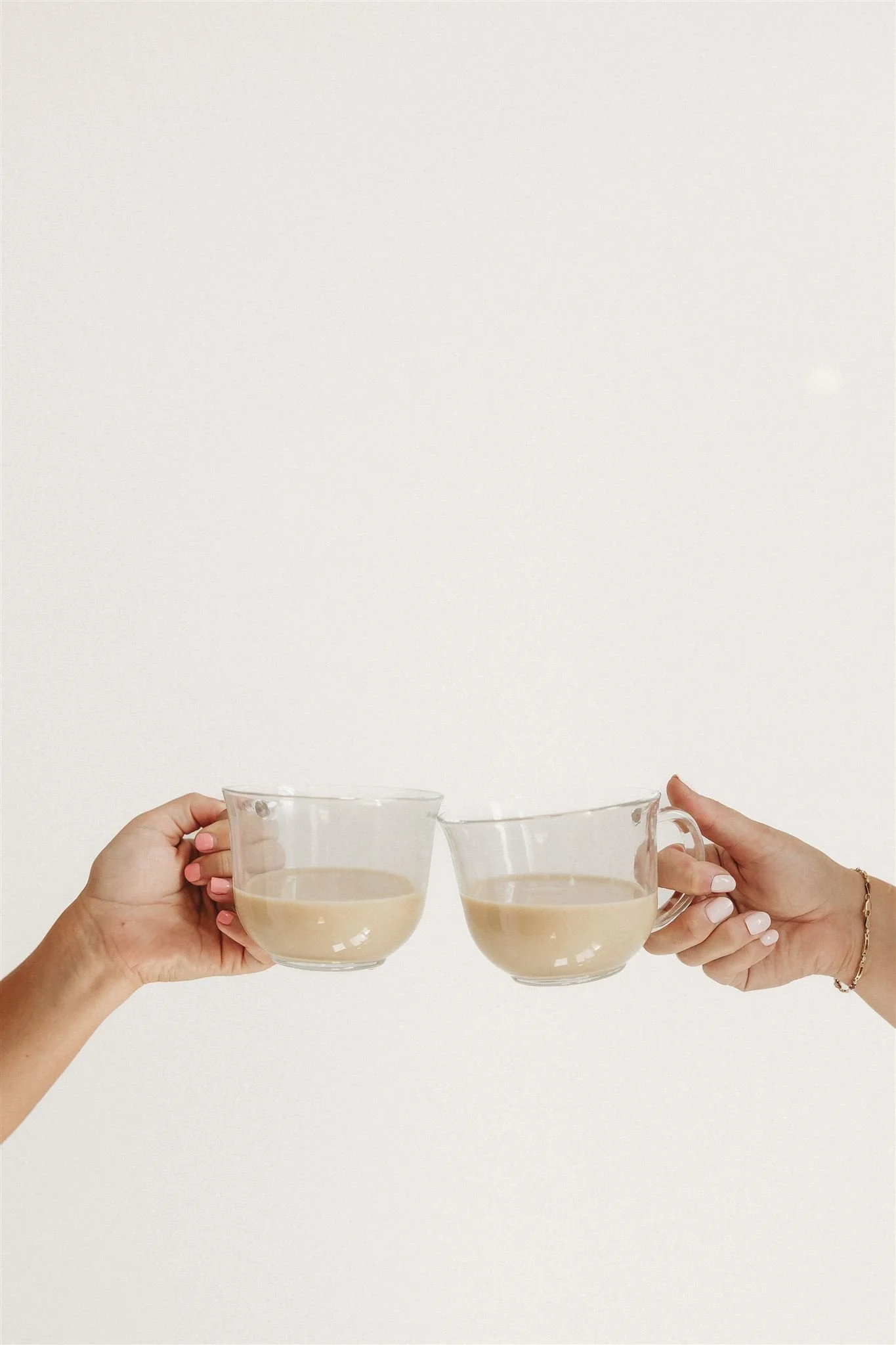 Two hands holding clear glass cups filled with coffee or tea, touching at the rims against a plain white background.