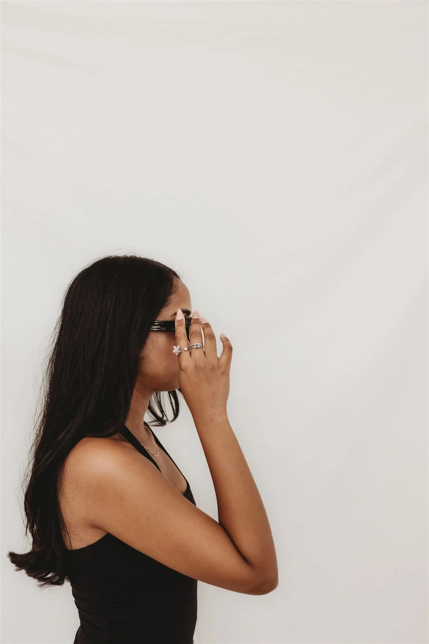 Woman with long dark hair wearing glasses, black tank top, and rings, covering her face with her hand, standing against a plain white wall.