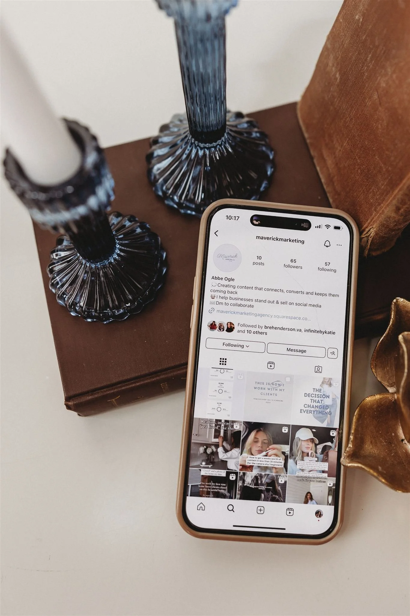 Smartphone displaying an Instagram profile on a white surface, surrounded by candle holders and decorative objects.