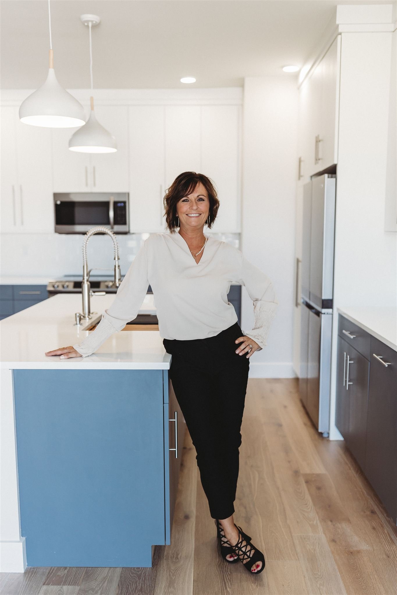 A woman with short brown hair wearing a white blouse and black pants posing in a modern kitchen.