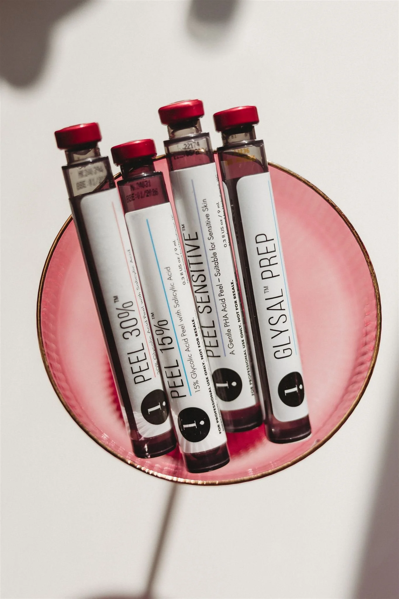 Four small glass vials with red caps, labeled 'PEEL 30%', 'PEEL 15%', 'PEEL SENSITIVE', and 'GEL SALT PREP', resting on a pink circular dish against a white background.