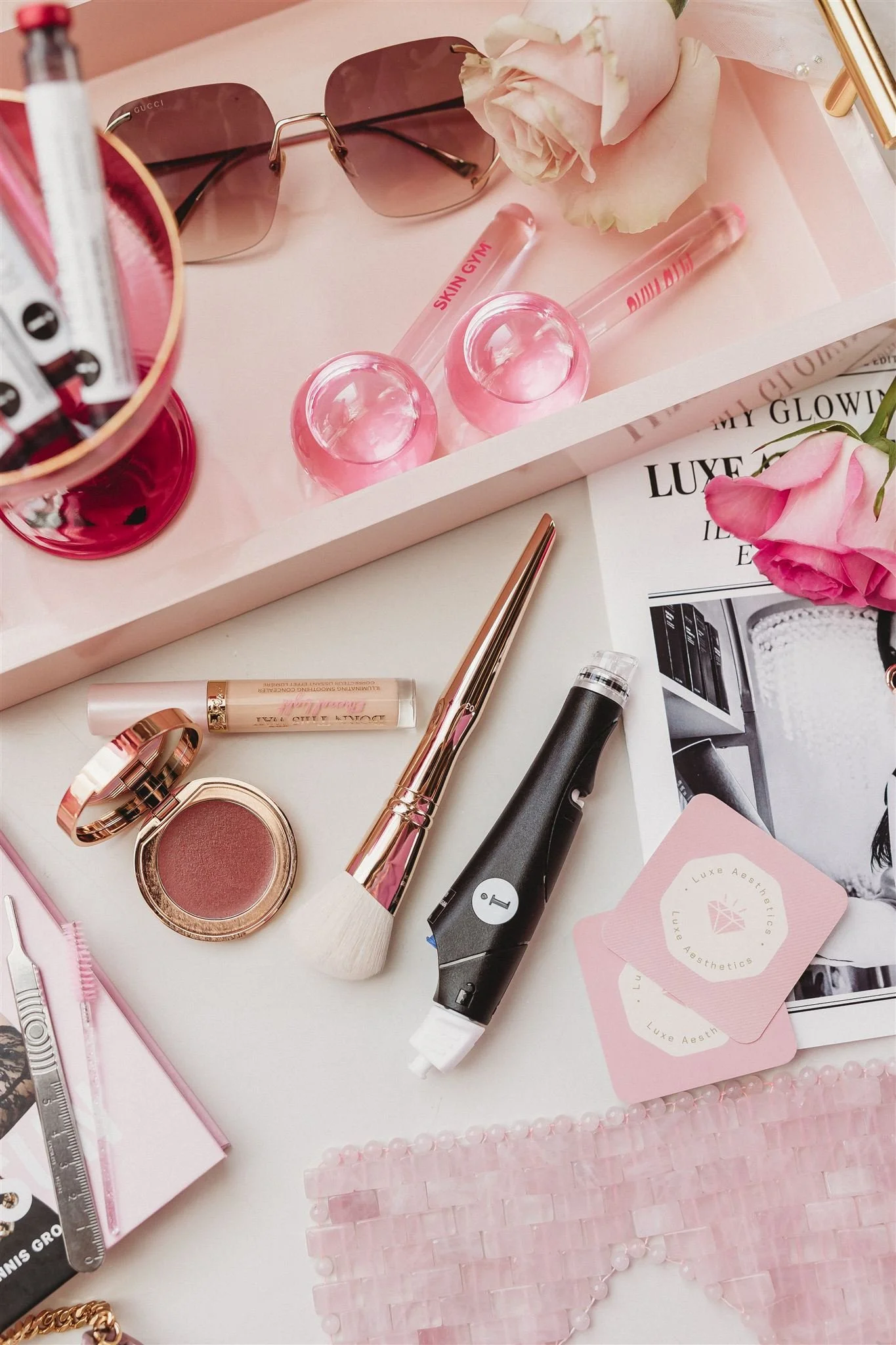 A flat lay of pink and black beauty and fashion accessories on a white surface, including sunglasses, pink roses, a tube of gloss, blush, a makeup brush, a facial device, and cards from Luxe Aesthetics.