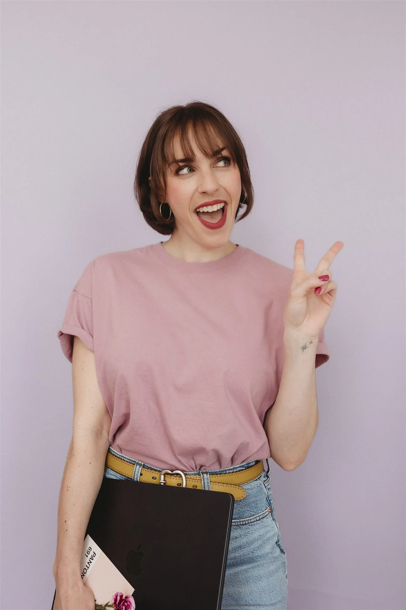 A young woman with short brown hair, wearing a pink t-shirt and jeans, making a peace sign with her right hand, holding a laptop and a magazine, smiling, against a light purple background.