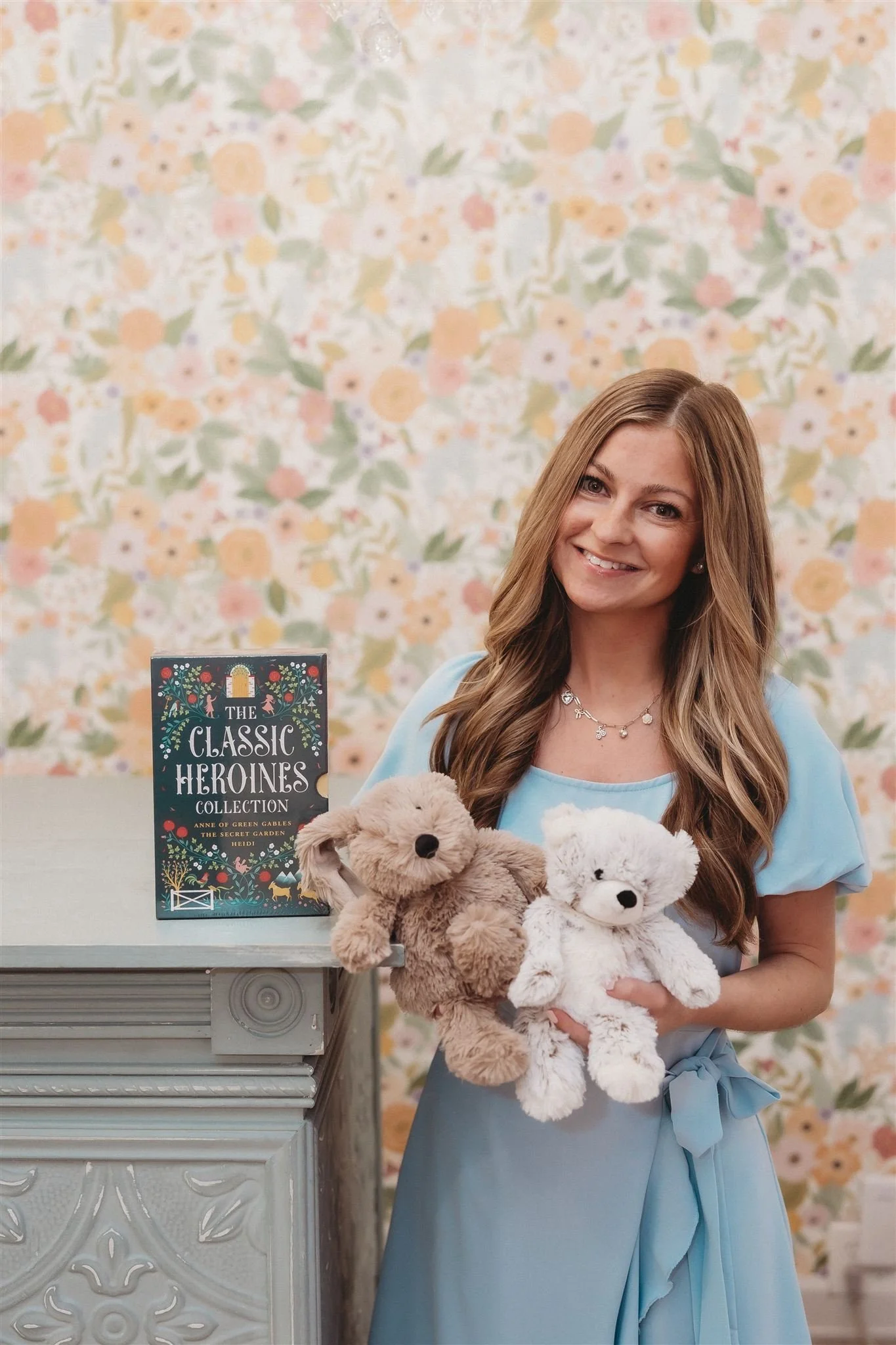 A smiling woman in a light blue dress holding two plush toys, a beige dog and a white teddy bear, standing next to a table with a book titled "The Classic Heroines Collection" against a floral patterned background.