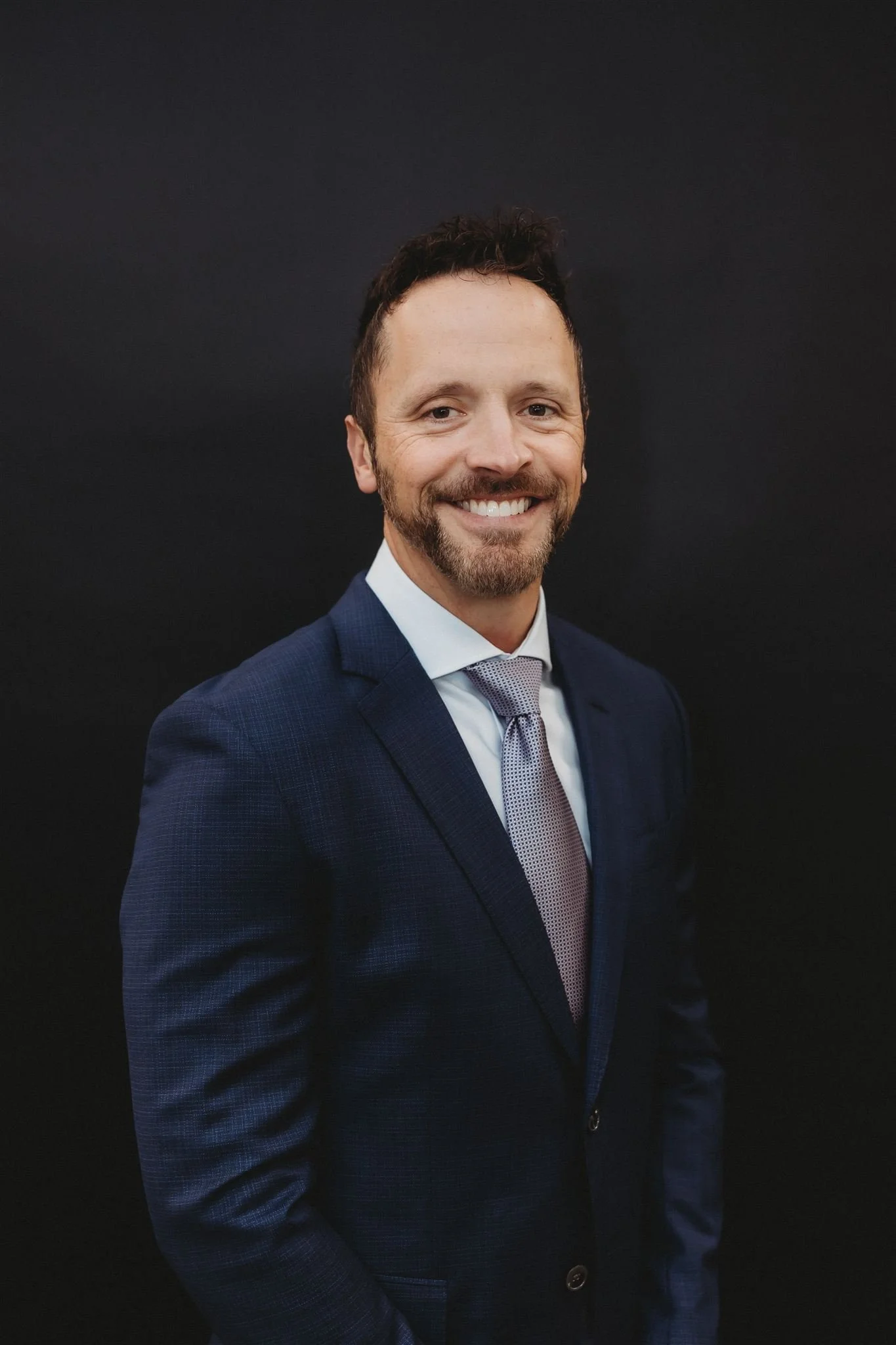A man in a dark blue suit with a light blue shirt and a light purple tie, smiling against a black background.