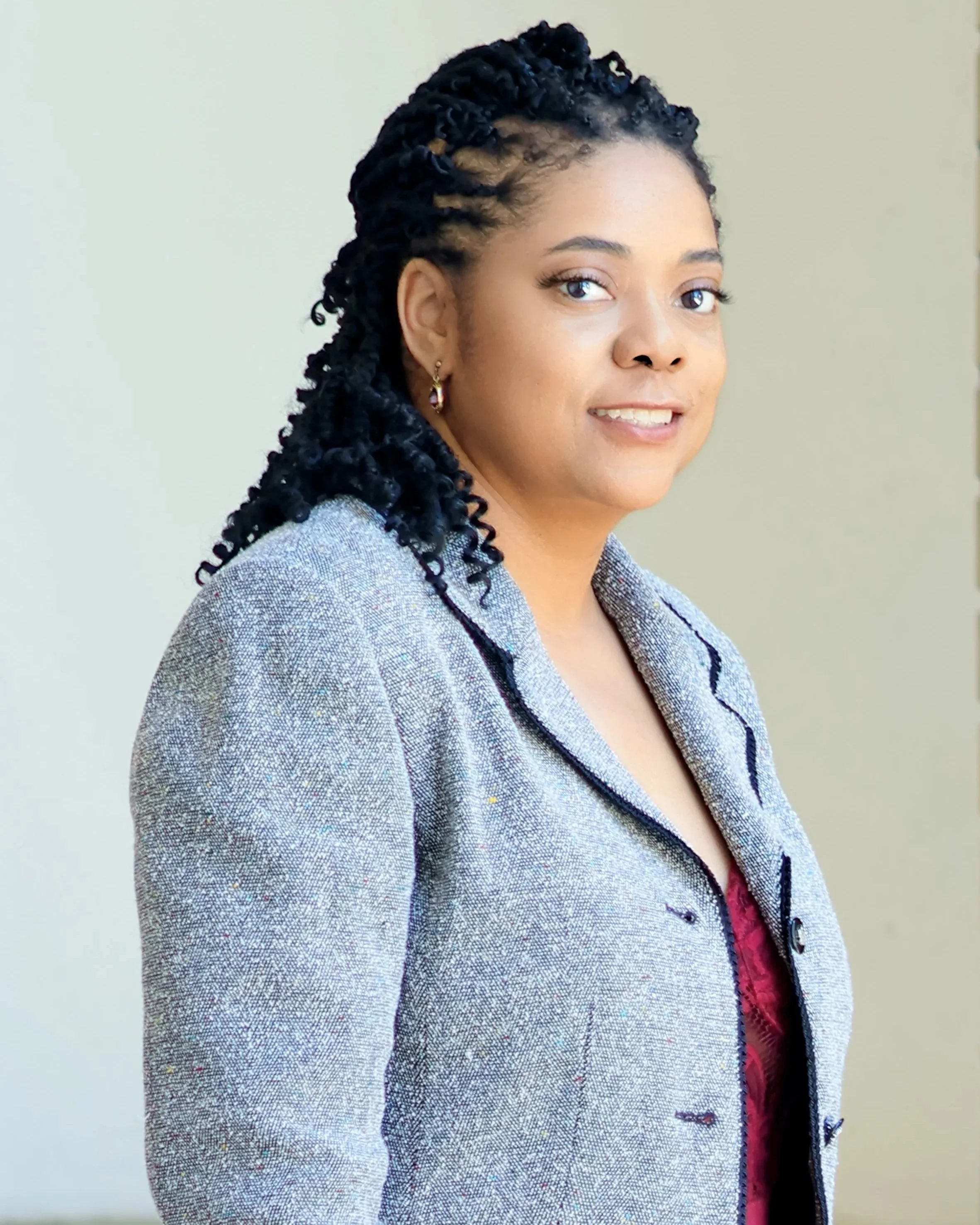 Young woman with medium skin tone, dark hair styled in twists, wearing earrings and a light-colored blazer, standing indoors with a neutral background.