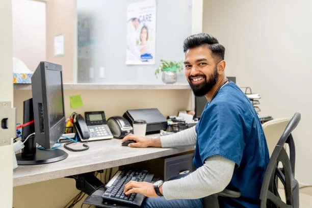 A smiling man in blue medical scrubs sitting at a desk in a healthcare office with a computer, phone, and medical supplies.