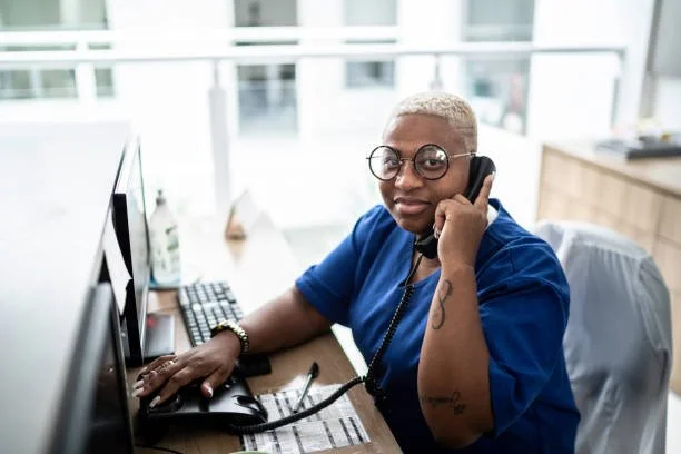 A woman with short blonde hair, glasses, and wearing blue scrubs, sits at a desk in an office, talking on the phone with a computer monitor and keyboard in front of her.