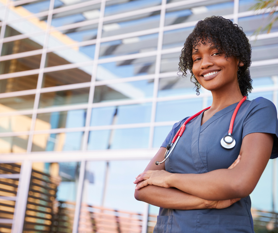 A smiling female healthcare professional wearing blue scrubs and a stethoscope standing outdoors in front of a modern glass building.