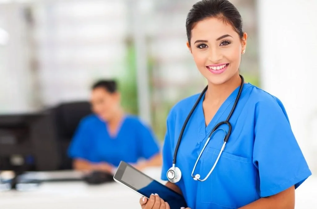 A smiling nurse in blue scrubs holding a tablet, with another nurse working at a computer in the background.