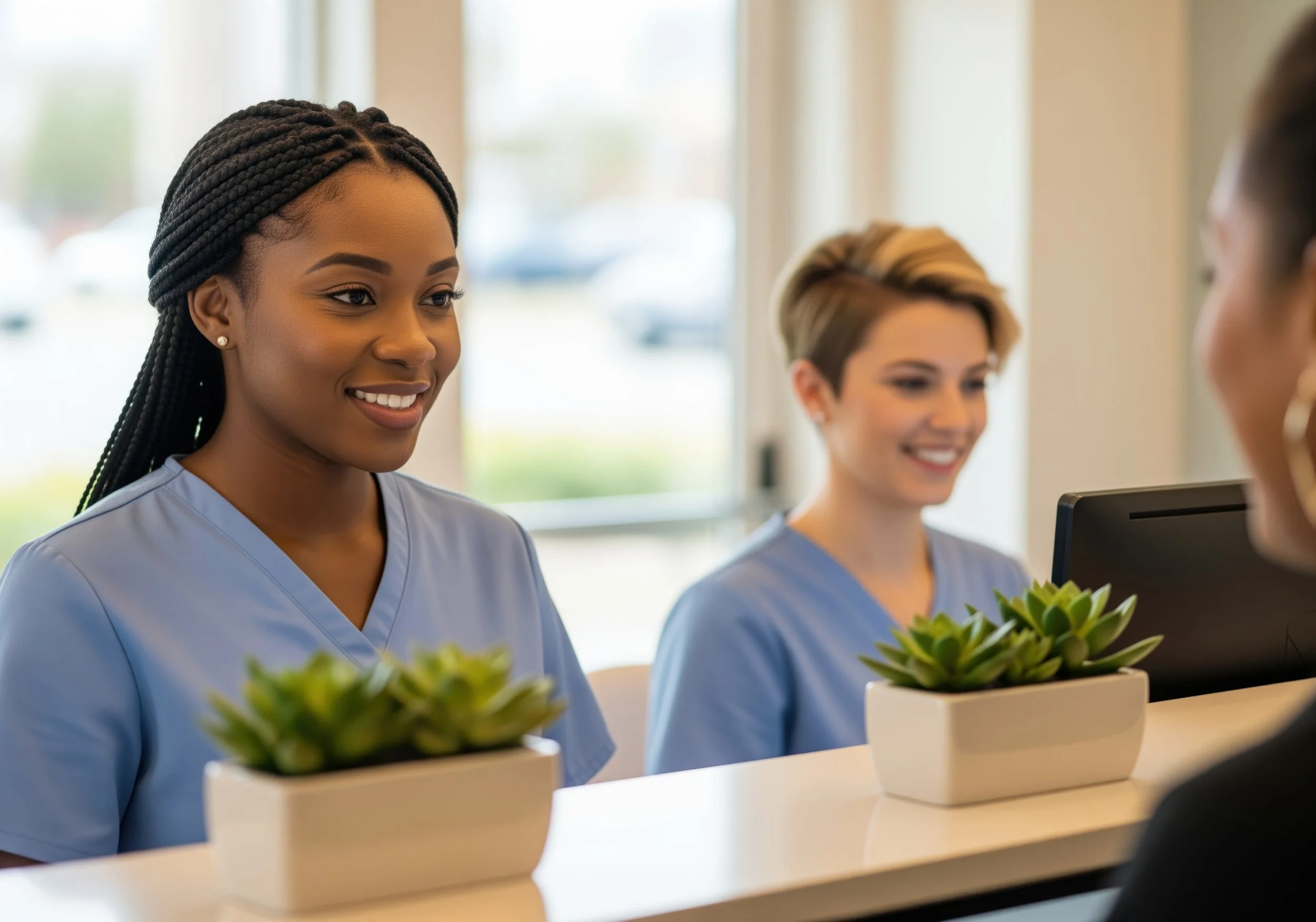 Two female healthcare workers in light blue scrubs handing a small potted succulent to a visitor at the reception desk. The reception area is bright with natural light.