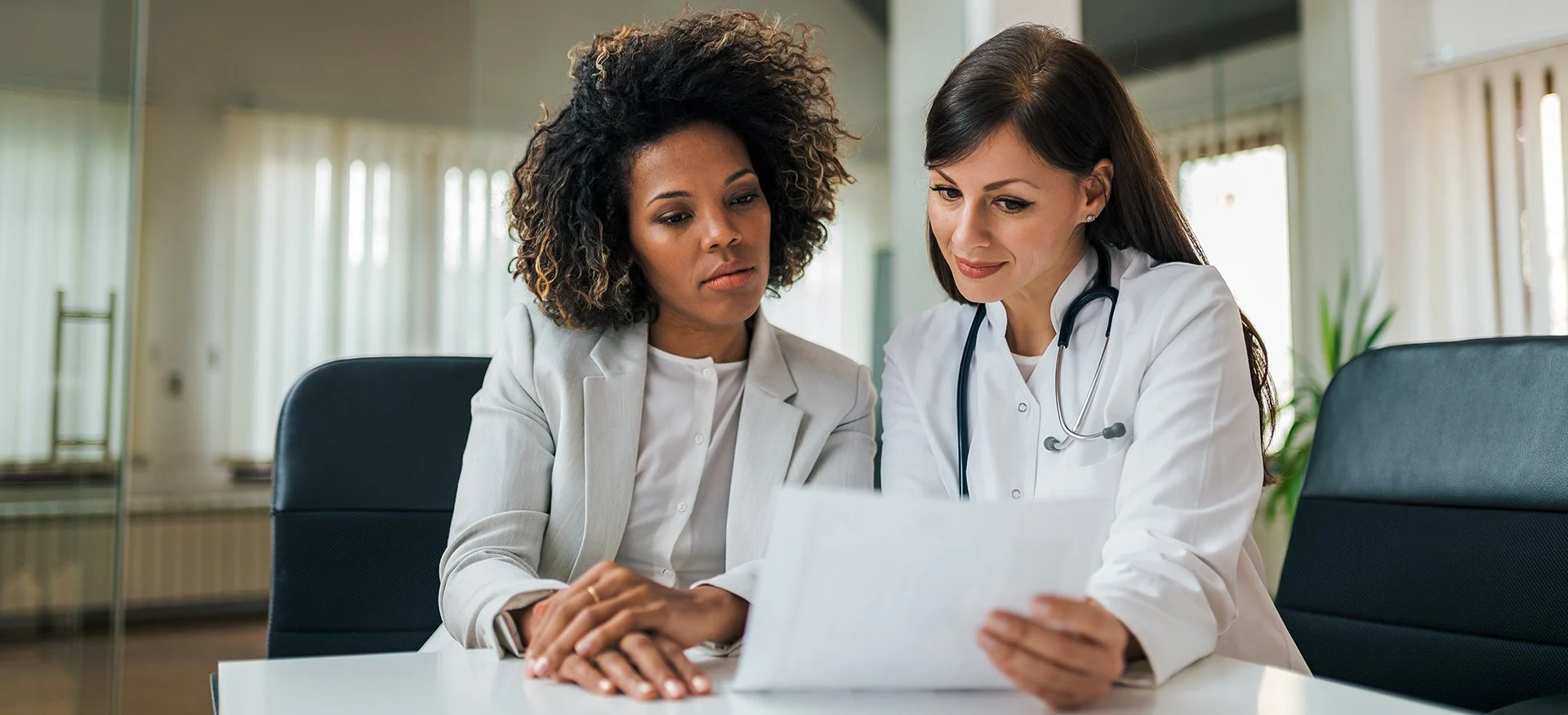 Two female healthcare professionals, one wearing a white coat and stethoscope, reviewing medical documents together in a consultation room.