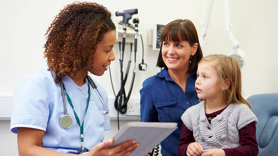 A young girl in a gray sweater talking to a female nurse with a stethoscope, and a woman smiling in a medical examination room.