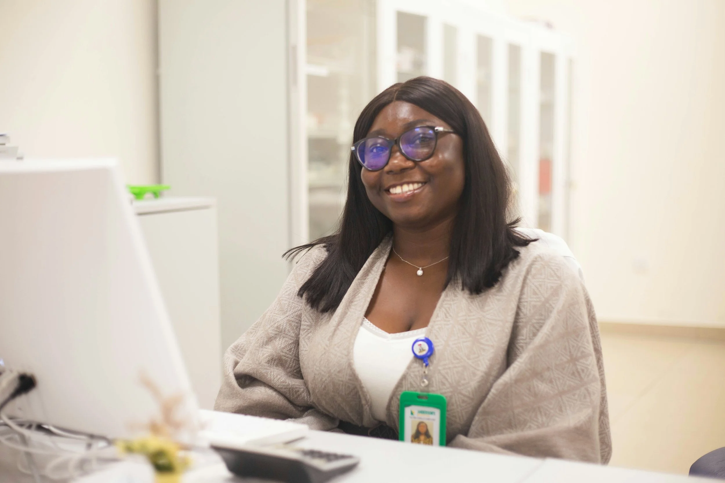A smiling woman with dark hair and glasses, wearing a beige jacket and an ID badge, working at a desk in an office.