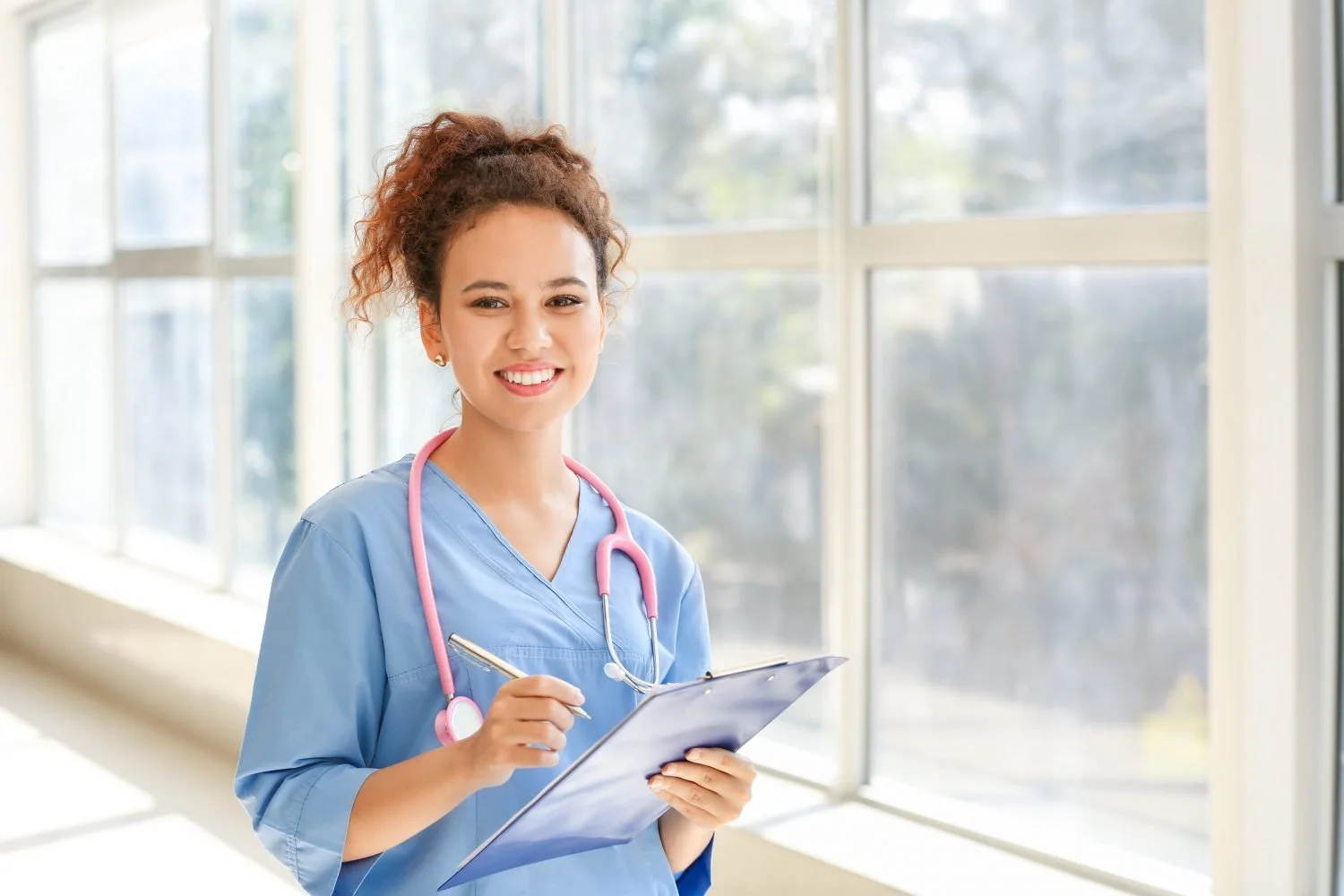 A young female healthcare professional in medical scrubs with a pink stethoscope around her neck, holding a clipboard and smiling at the camera inside a bright hospital corridor with large windows.