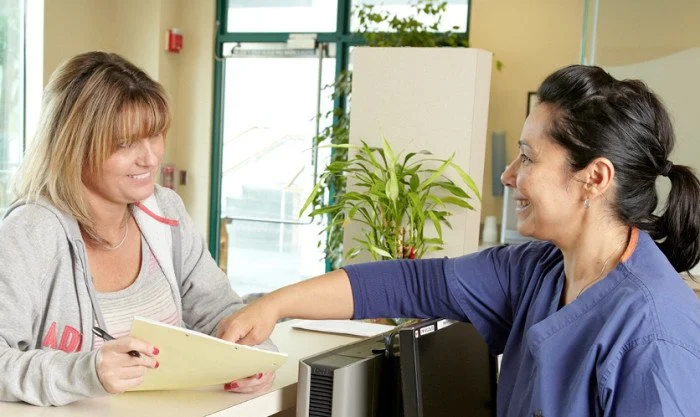 A woman sitting at a reception desk shaking hands with a healthcare worker in scrubs