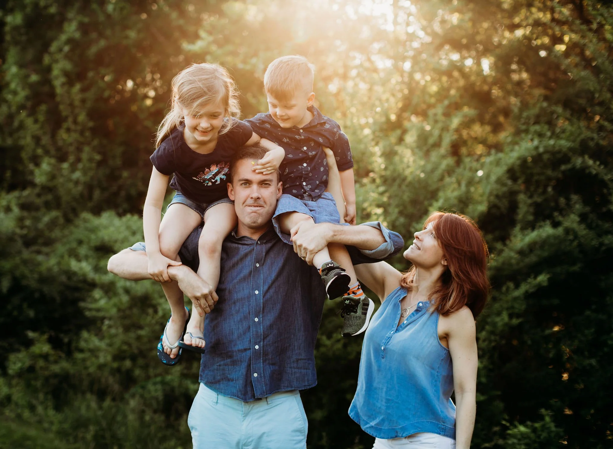 Family outdoors with a man holding two children on his shoulders and a woman standing beside them, all smiling in a green park at sunset.