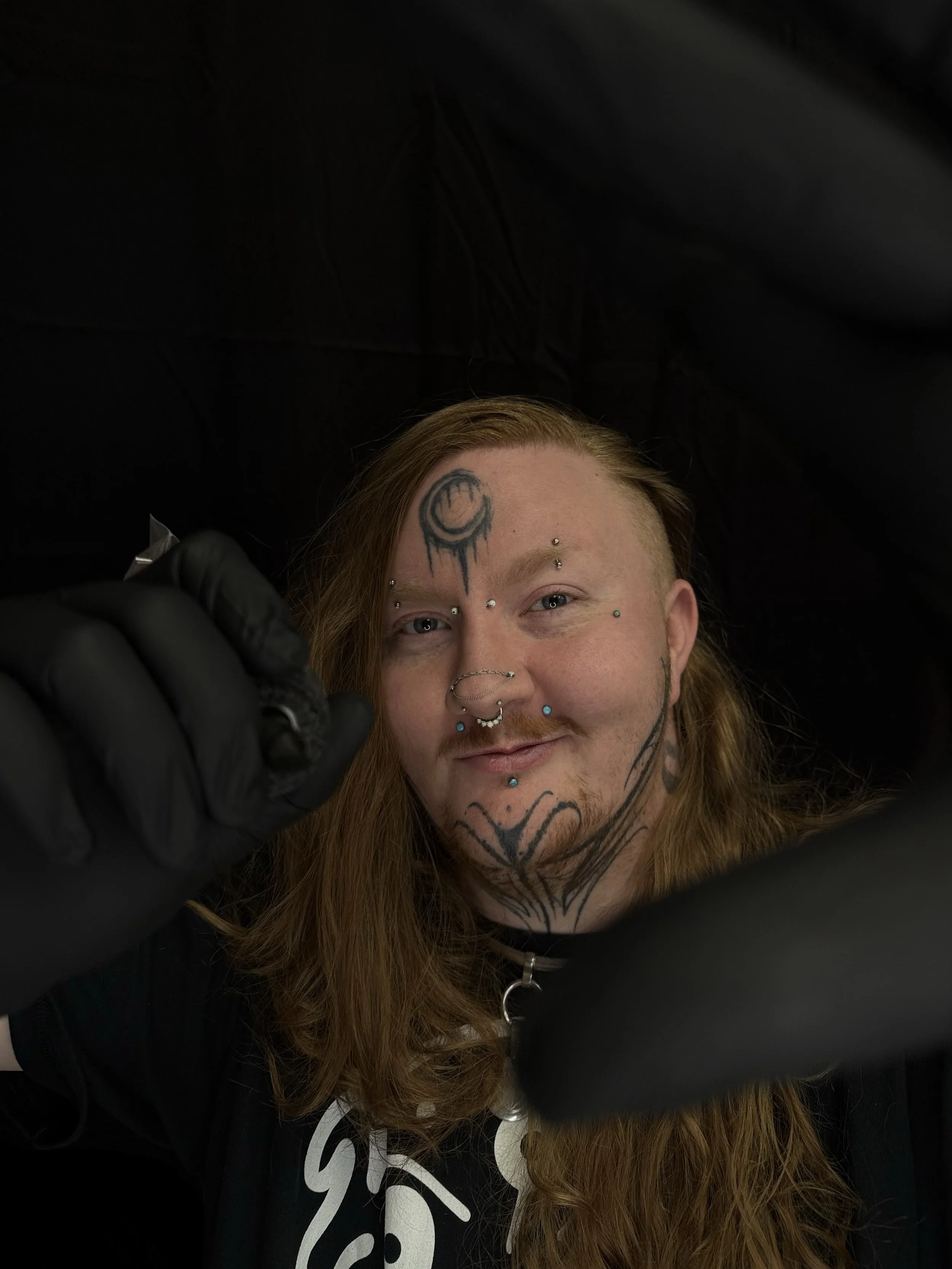 Person with long red hair and facial tattoos, smiling, lying down, wearing black gloves, holding a tattoo machine. Tattoo Artist from Parachute, Colorado at Sad Circus Studio.