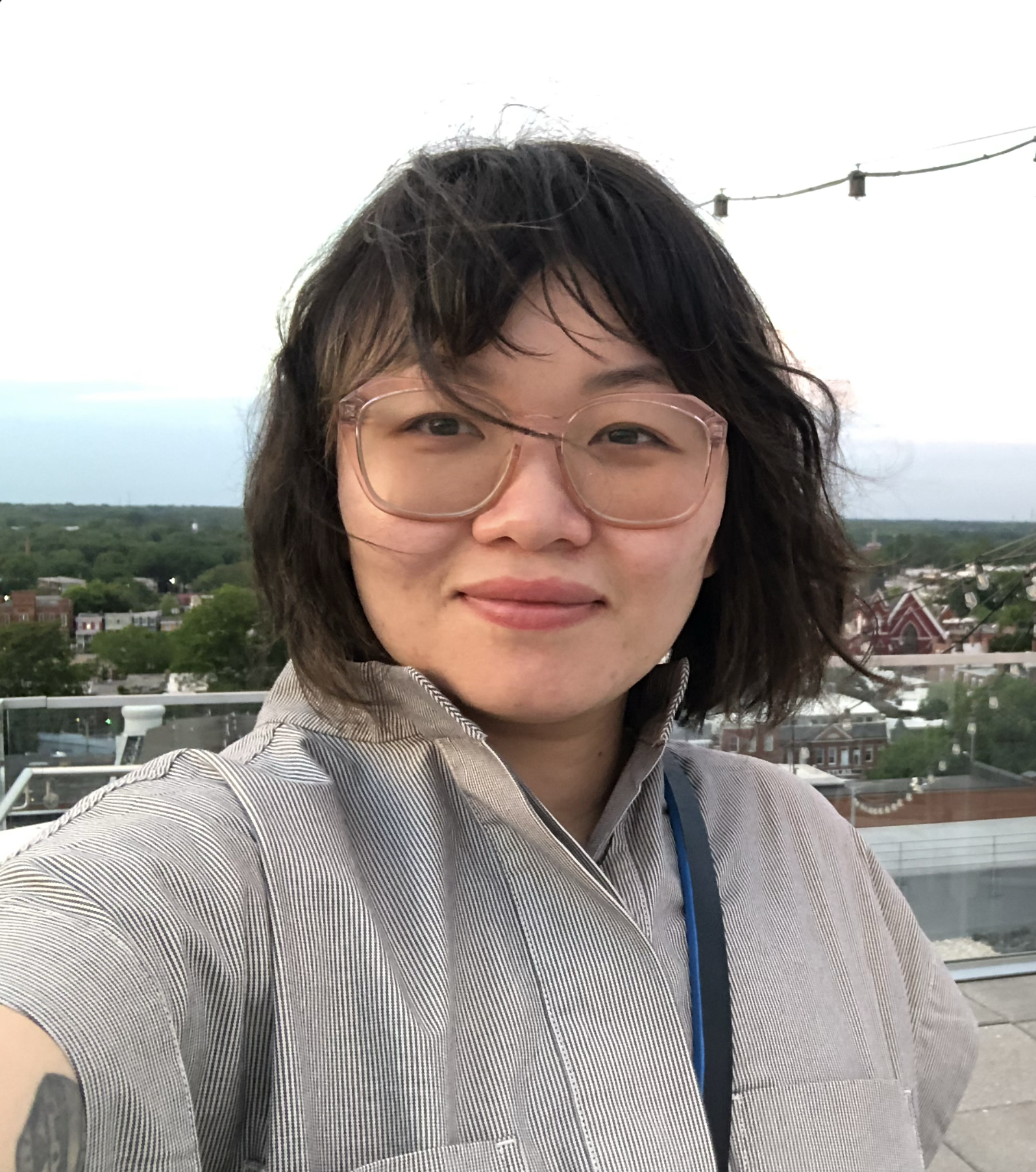A woman with glasses taking a selfie on a rooftop. She is wearing a striped shirt. The background features a cityscape with roofs, trees, and power lines under a cloudy sky.