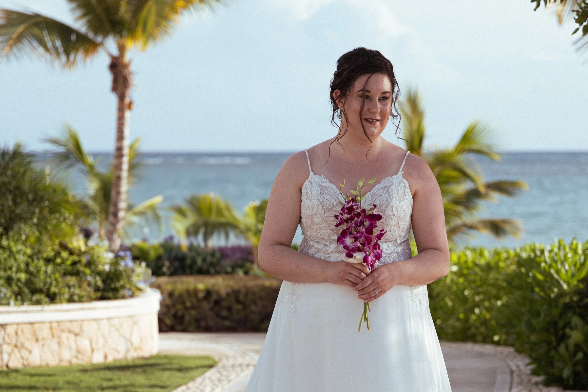 A woman in a white lace dress holding a bouquet of purple and white flowers, standing outdoors near the ocean with palm trees and greenery around.