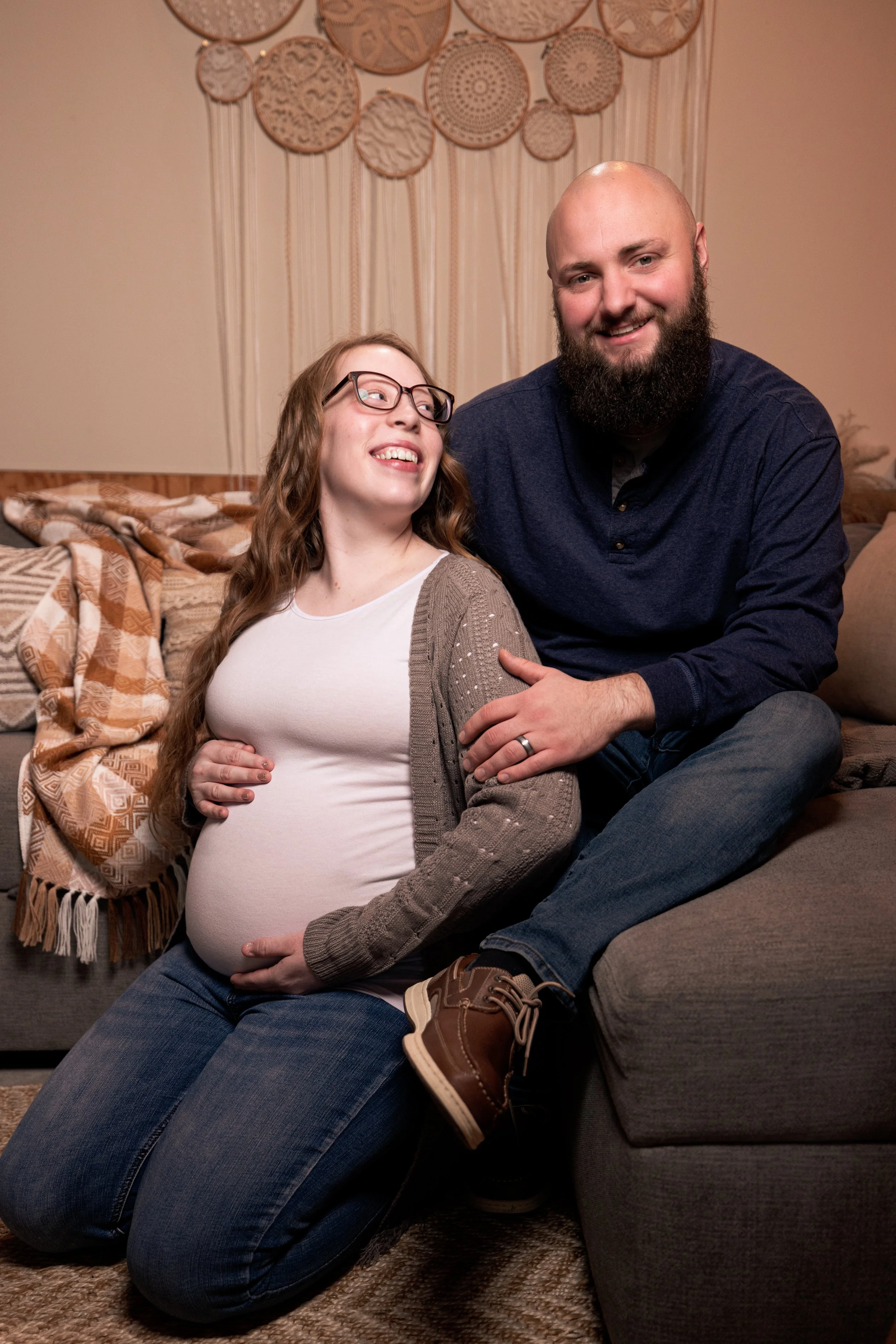 A pregnant woman with long brown wavy hair and glasses sitting on her knees on the floor, smiling and looking at a man with a beard, who is sitting on the sofa next to her. The man is wearing a dark blue shirt and jeans, smiling, and has an arm around the woman. They are in a cozy living room with a decorative wall hanging behind them, a patterned blanket on the sofa, and a textured rug on the floor.