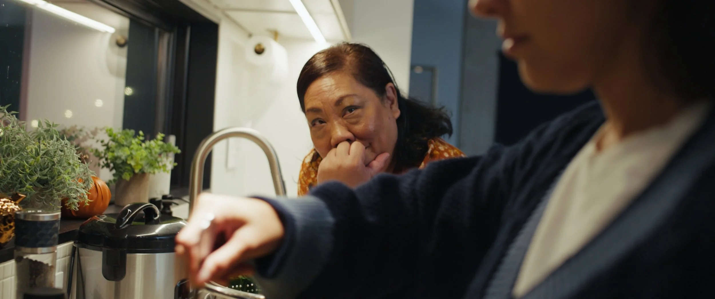 A woman with dark hair and wearing an orange patterned top looking at a young person in a dark sweater, who is pouring something into a container in a kitchen decorated with potted plants.