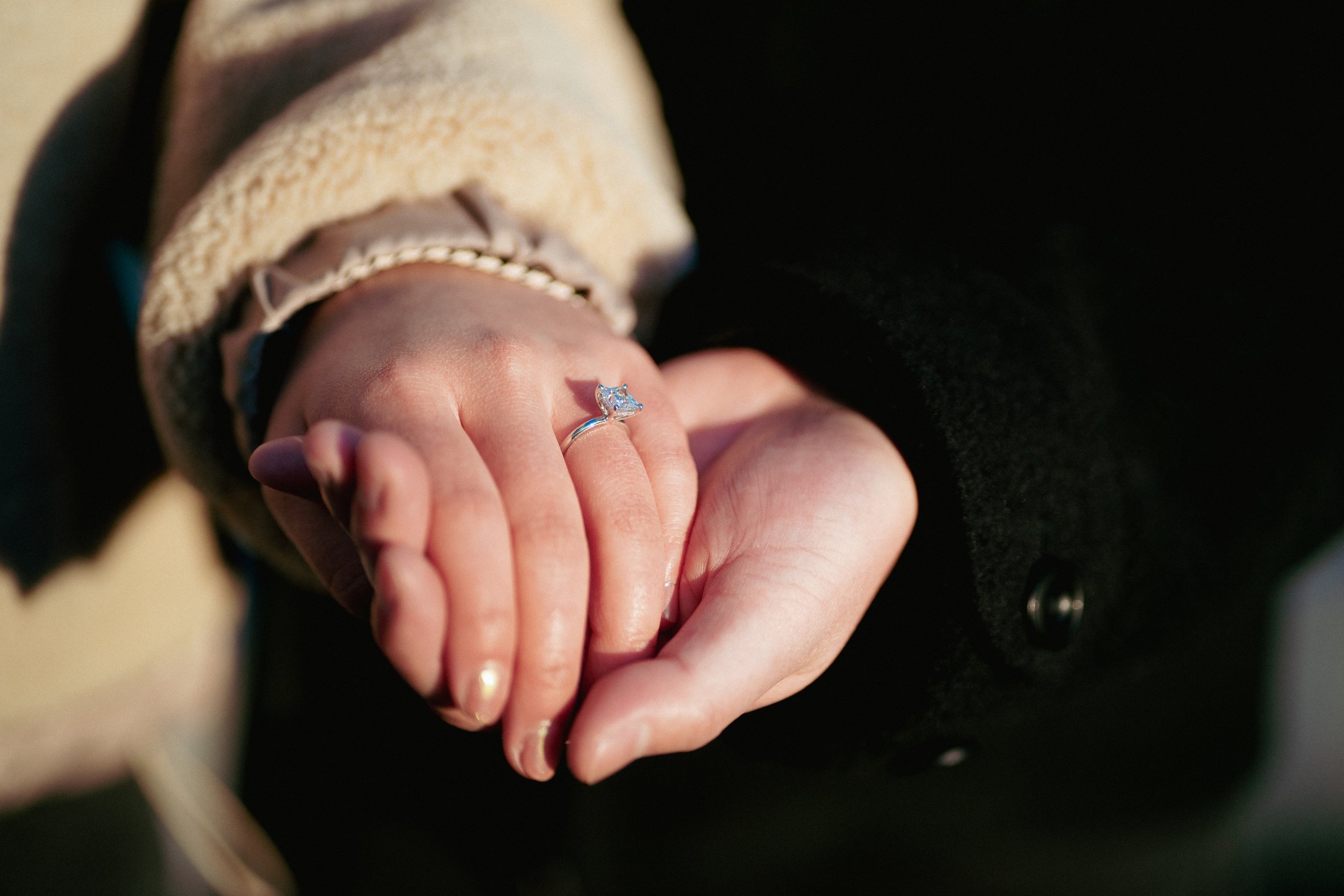Close-up of a person's hand with an engagement ring featuring a large diamond, with the person wearing a beige and black outfit.