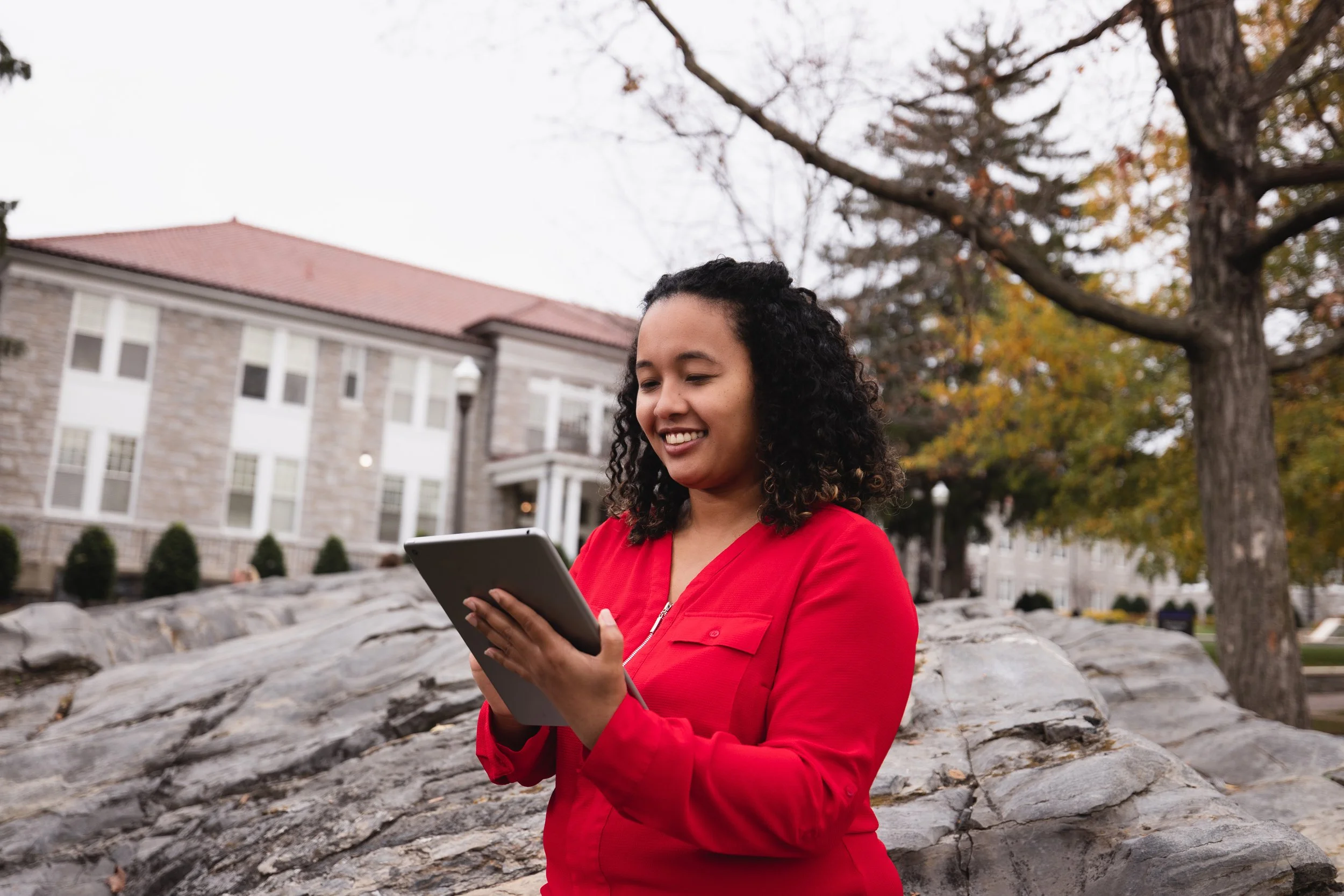 A woman with curly hair wearing a red jacket, smiling and looking at a tablet, standing outdoors on a stone pathway with trees and a building in the background during autumn.