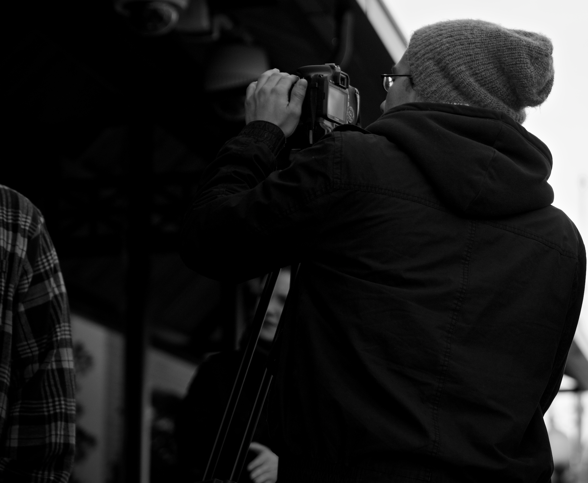 A person wearing a beanie and glasses, holding a camera as if taking a photograph, standing near shelves in what appears to be a train station. The image is in black and white.