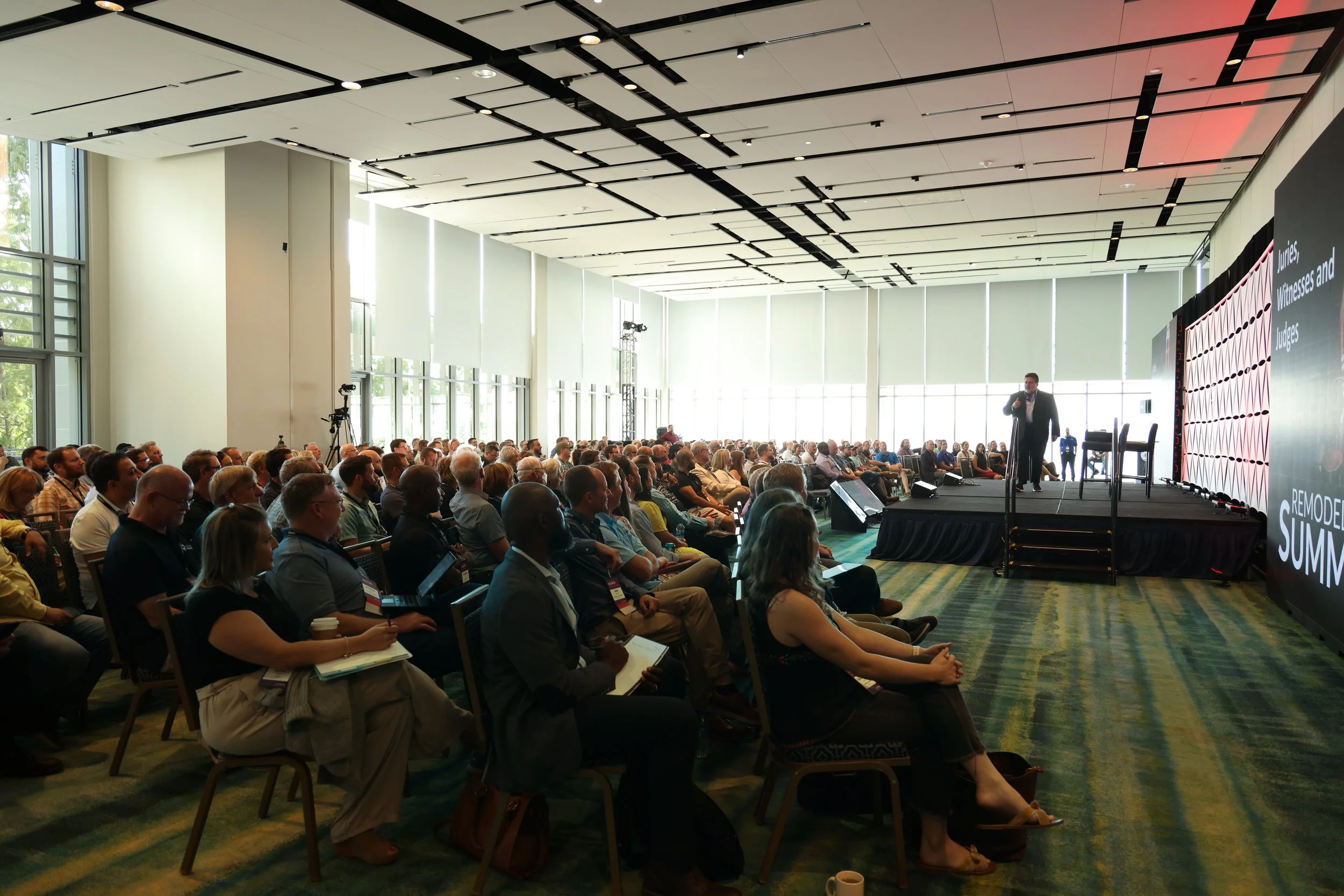 A large conference room filled with seated attendees listening to a speaker on stage. The ceiling has modern black linear lighting fixtures, and the room has large windows with white blinds letting in natural light. On the stage, a person is speaking into a microphone with a large screen displaying text behind them.