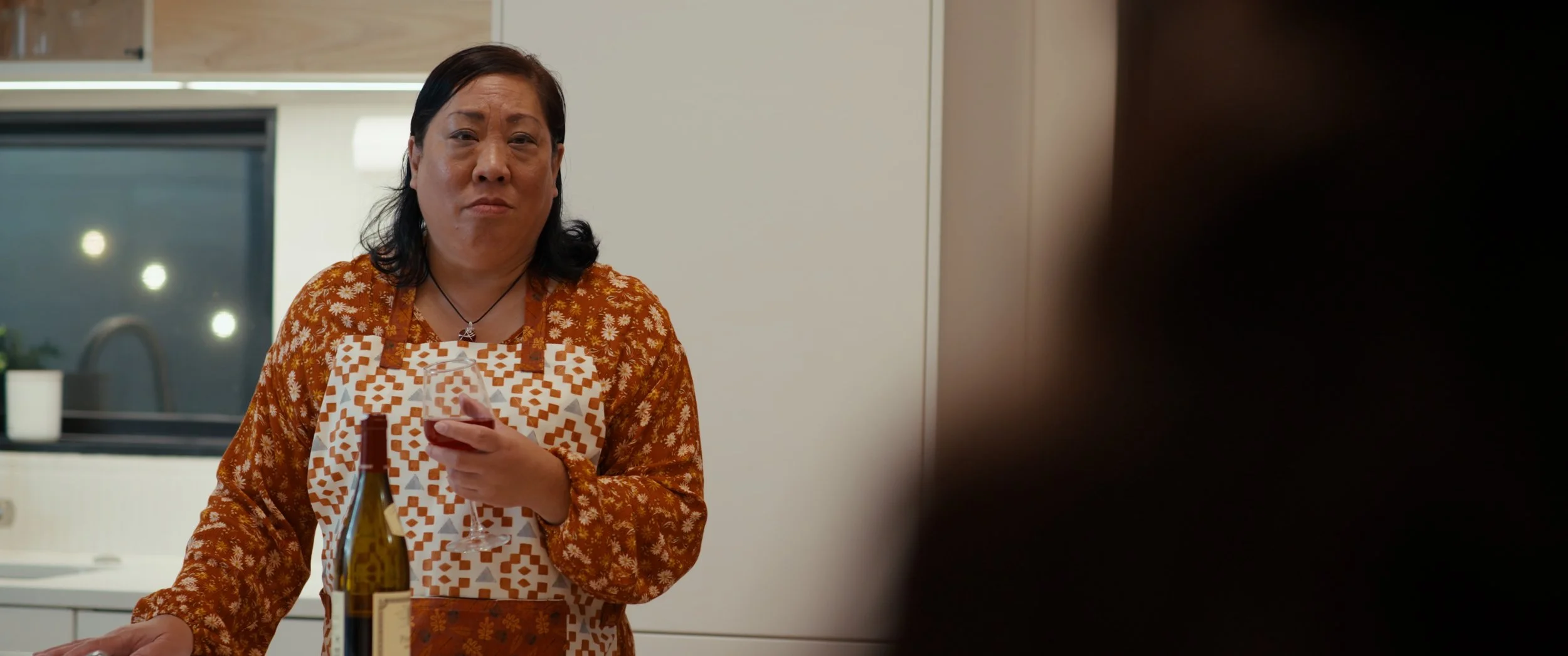 A woman in a floral apron holding a glass of red wine, standing in a modern kitchen.