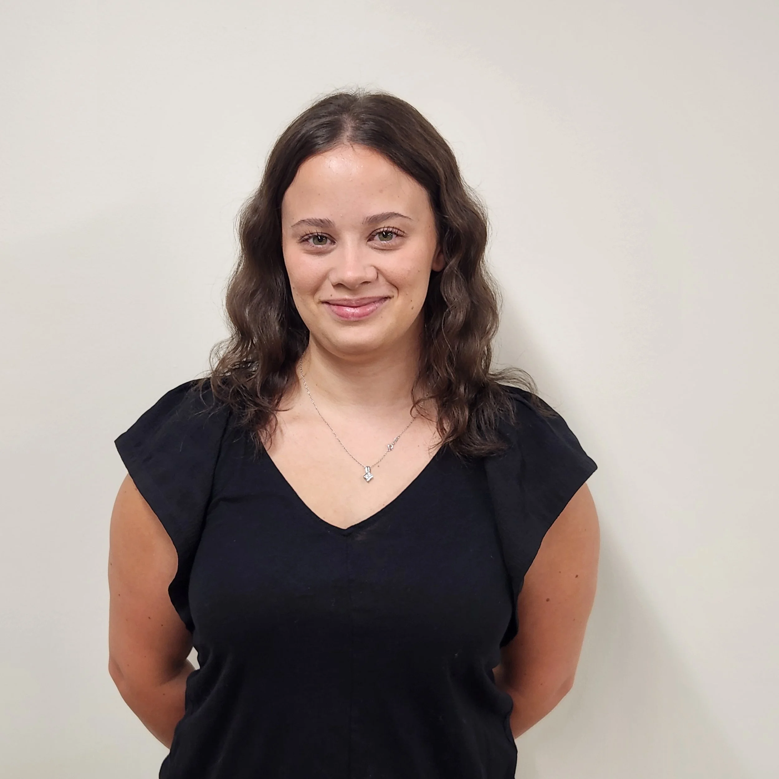 A woman with curly brown hair, light skin, and wearing a black top, smiling against a plain light-colored wall.