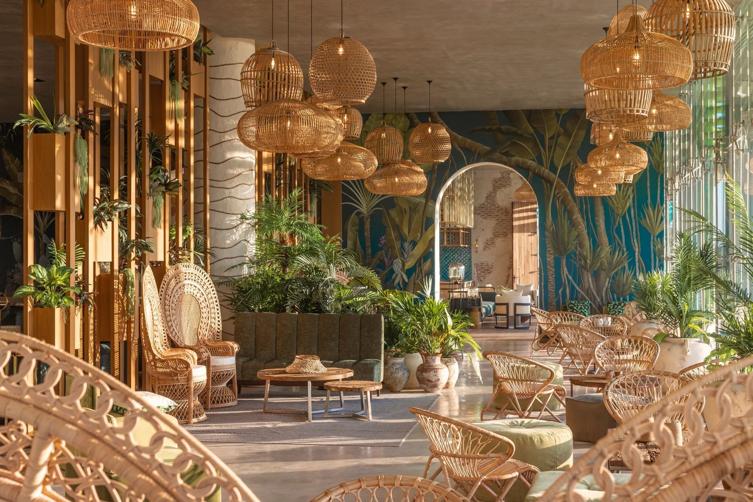 Tropical-themed interior with wicker furniture, hanging woven light fixtures, green plants, and a mural wall. Features a mix of chairs, tables, and a large archway leading to another room.