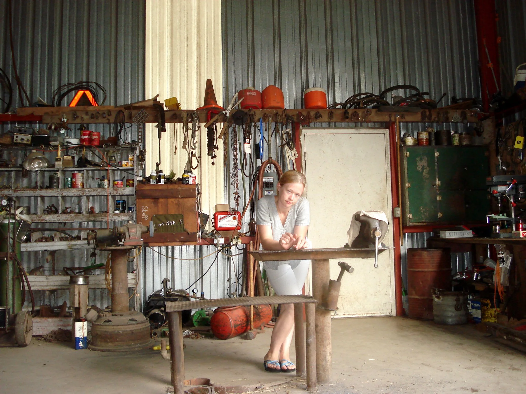 A young woman with blonde hair in a ponytail, wearing a gray shirt and flip-flops, sitting on a workbench in a cluttered barn with tools, storage bins, and equipment hanging on the walls.
