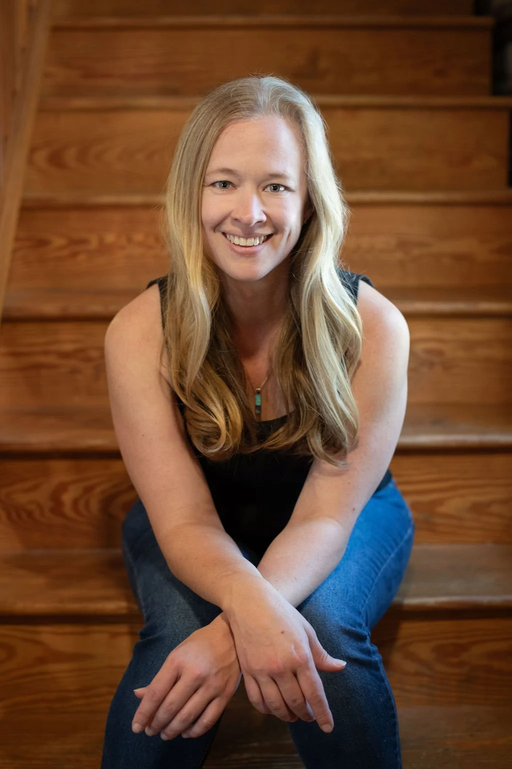 A woman with long blonde hair, smiling, sitting on wooden stairs, wearing a black sleeveless top and blue jeans.