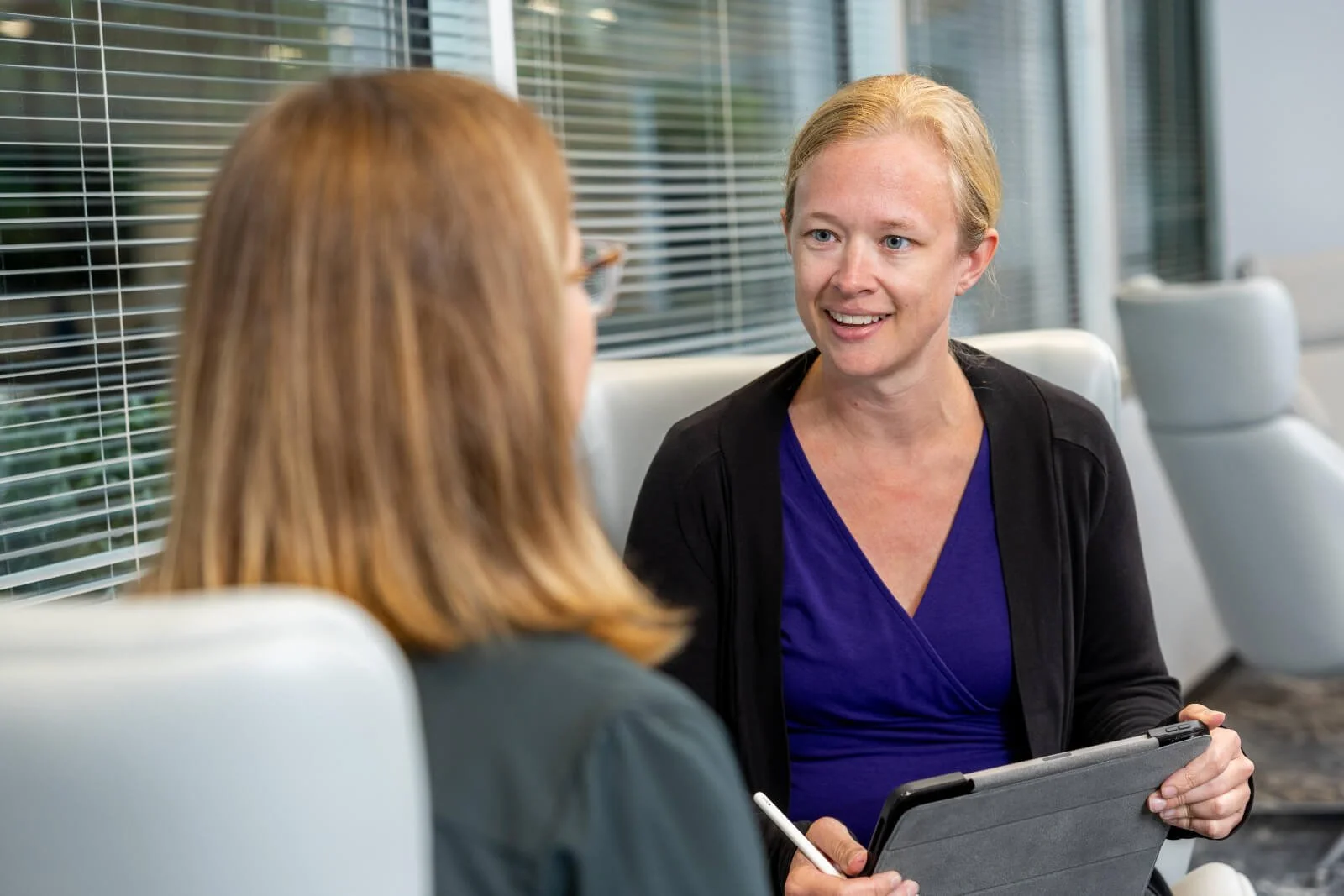 Two women engaged in conversation in a modern office or conference room, with a woman holding a tablet and a stylus.
