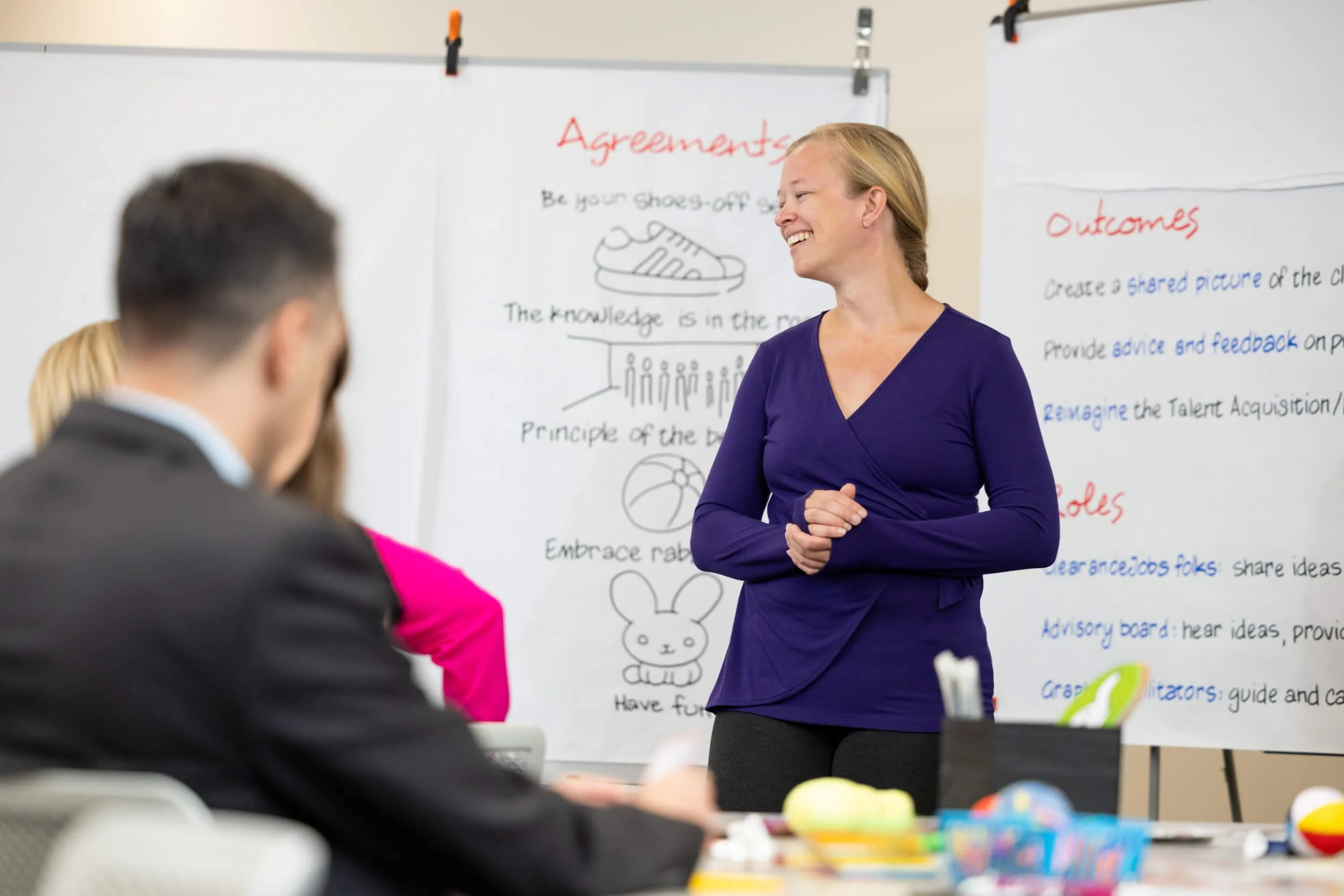 A woman with blonde hair smiling and standing in front of a whiteboard with notes, drawings, and text, teaching a class. Several people are seated in the foreground, with only parts of their heads visible.